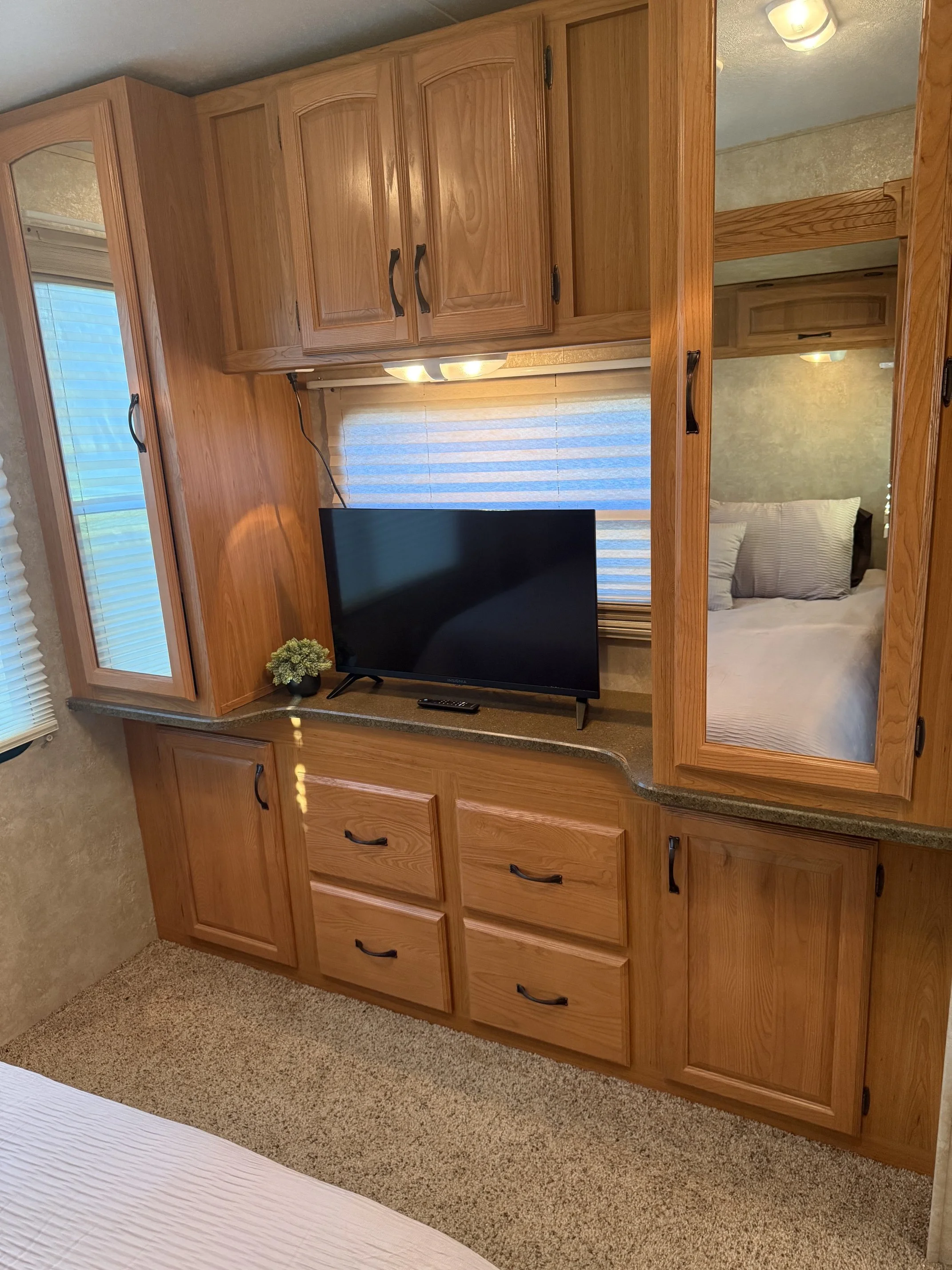 Interior of a room with a wooden entertainment center, a flat-screen TV, a small potted plant, a window with blinds, and a bed with pillows visible through a mirror and doorway.