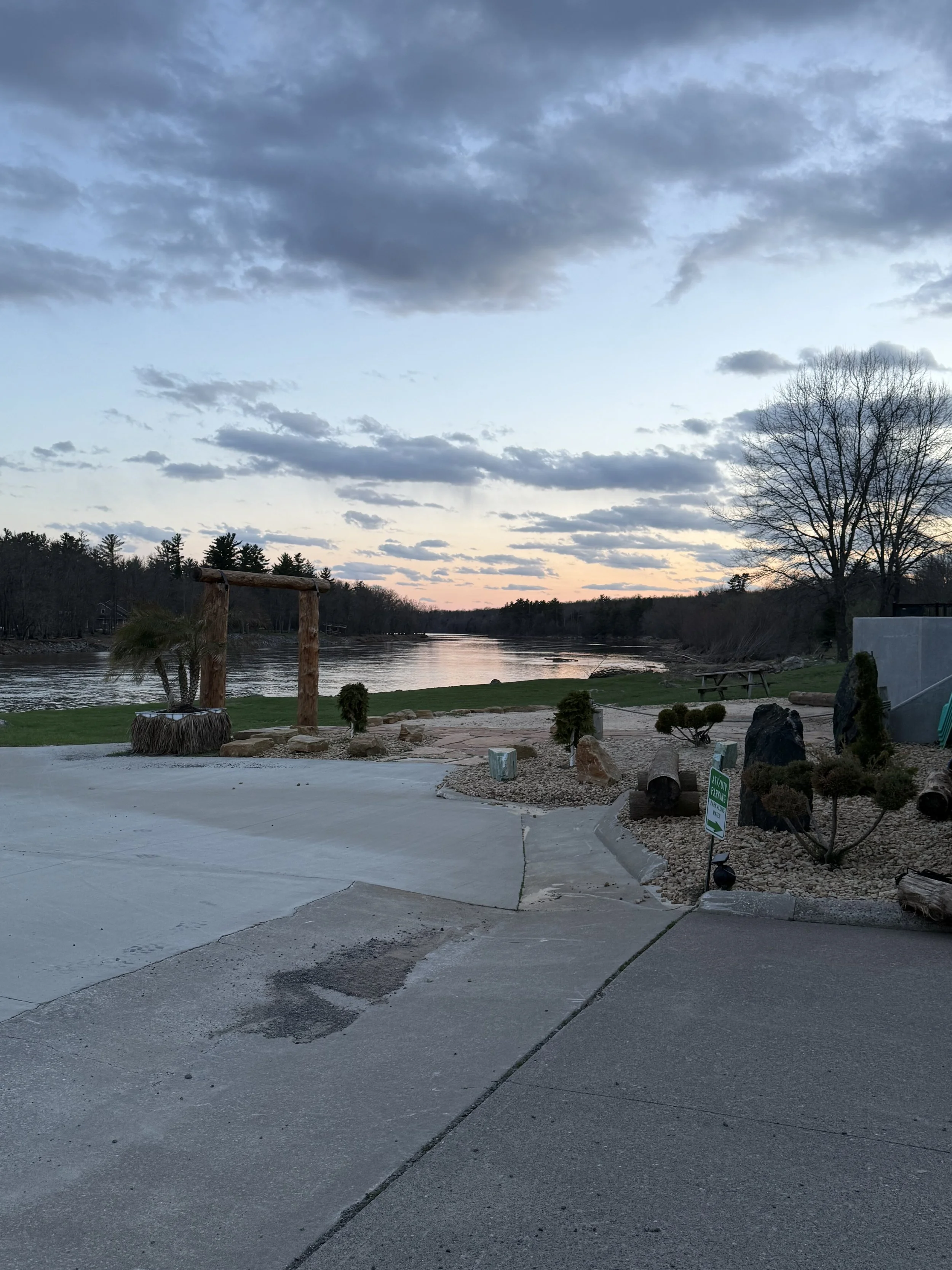 A river flowing through a landscape at dusk with a cloudy sky. There is a wooden structure near the riverbank, some bushes, rocks, and a park sign. Trees are visible in the background.