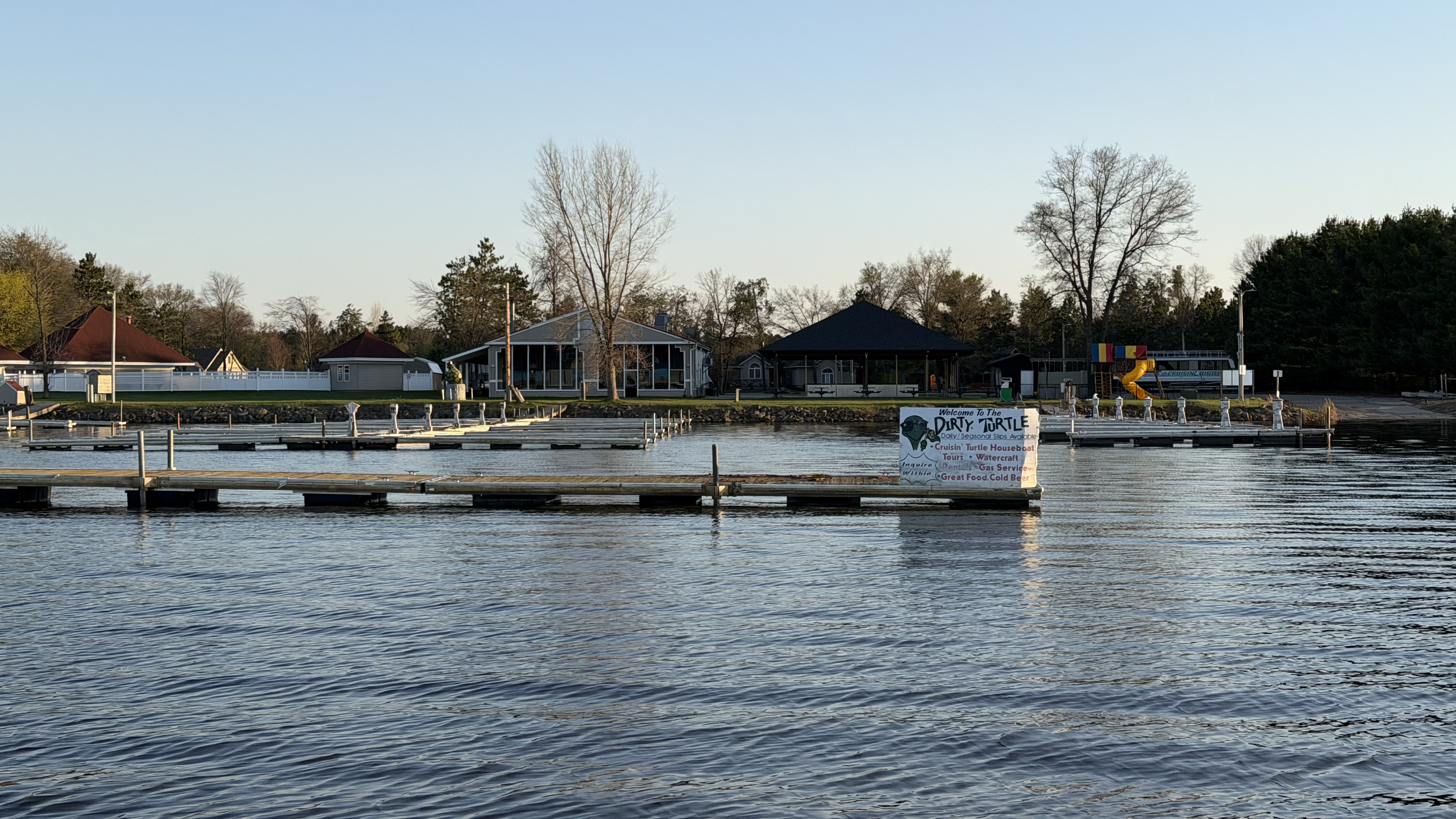 A waterfront scene with a dock, trees, and buildings, including a sign advertising a turtle houseboat tour and watercraft rentals.
