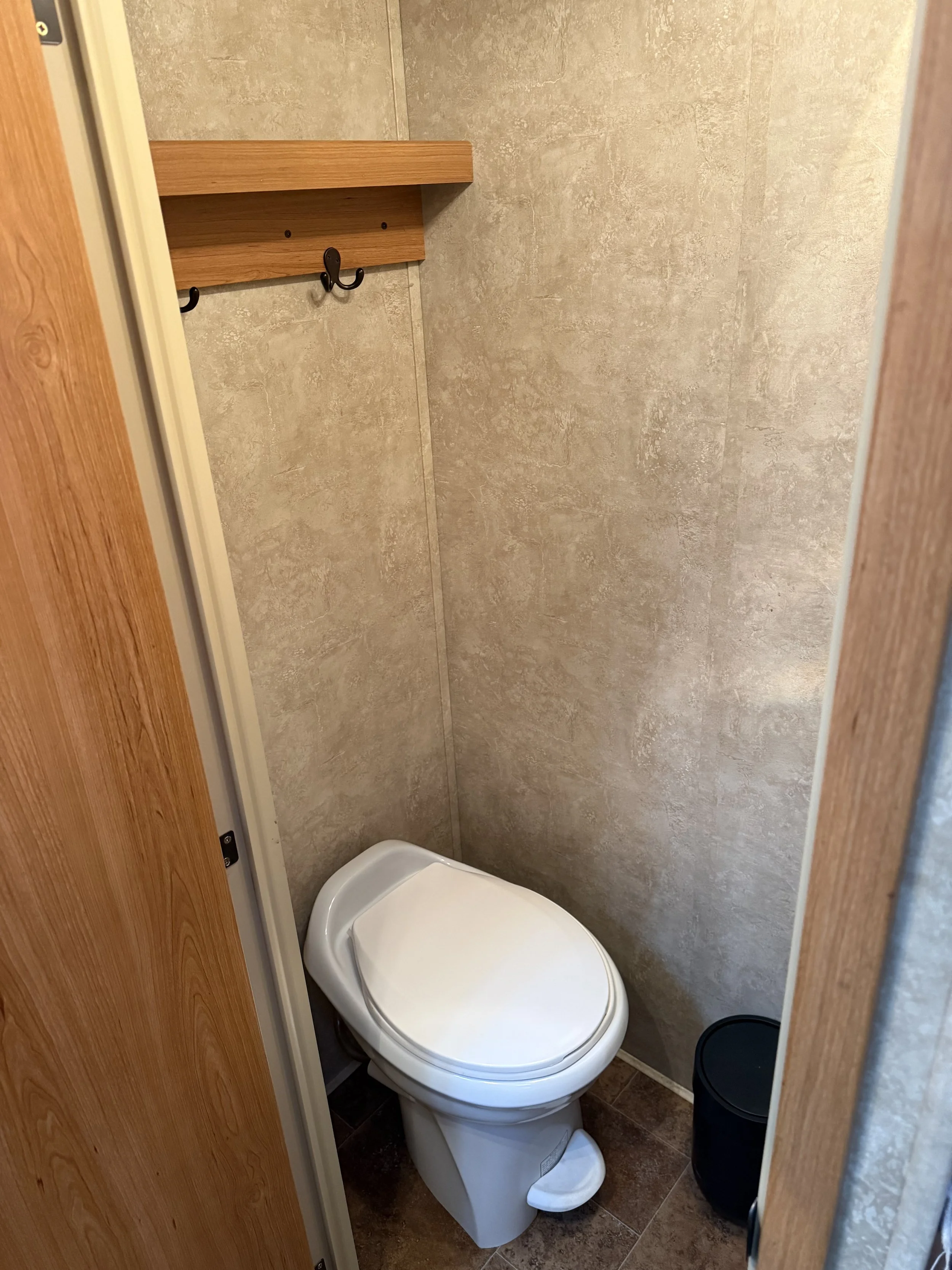 Small bathroom stall with a white toilet, a black trash bin, a wooden shelf with hooks, and beige walls.