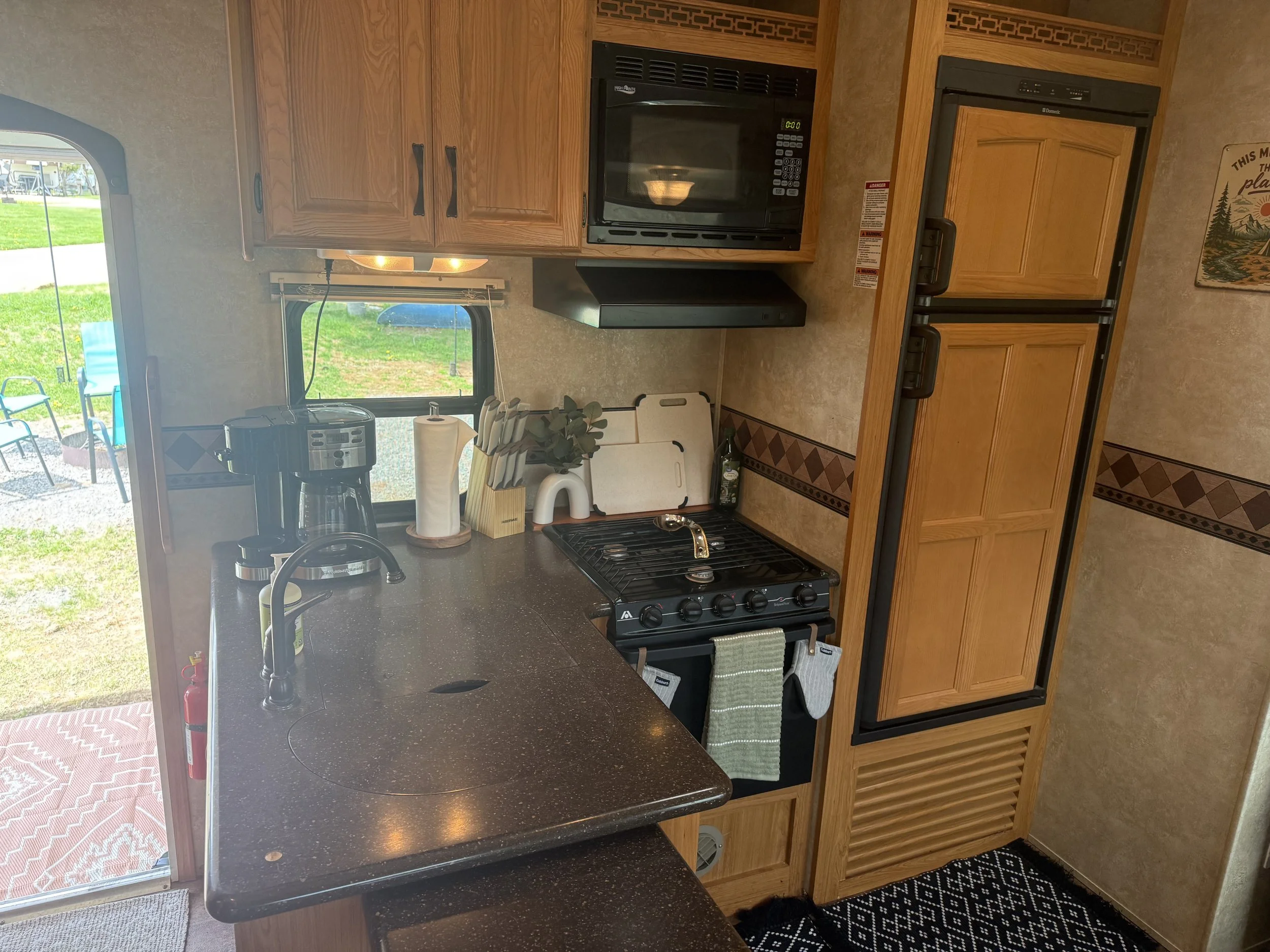 Interior of a camper kitchen with wooden cabinets, a microwave, a refrigerator, a small stove, a sink, and a window showing an outdoor camping area with chairs and grass.