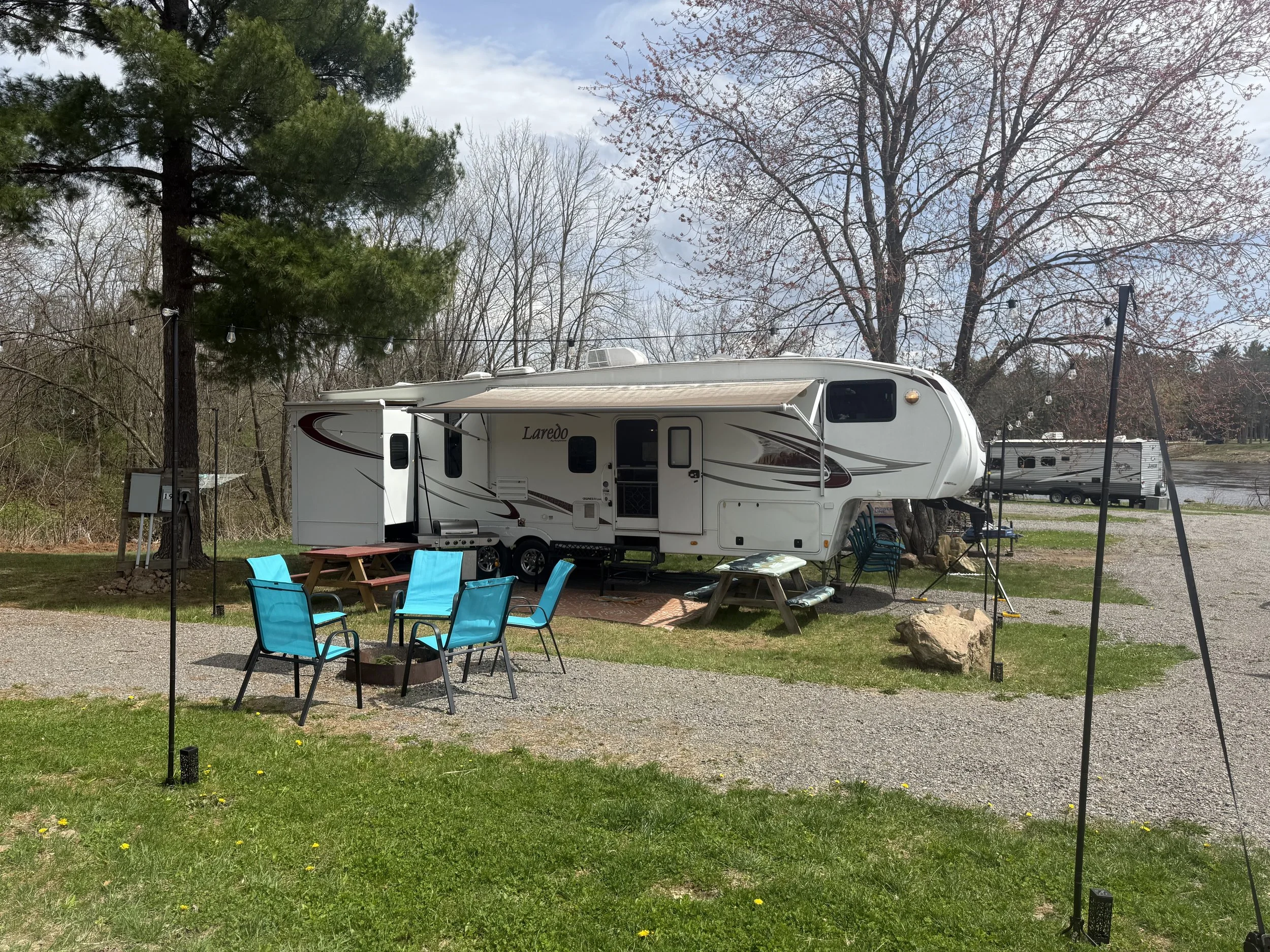 A white travel trailer with the name 'Laredo' written on it, parked outdoors under trees with string lights hanging above. There is a grassy area with blue chairs and a wooden picnic table in front of the trailer.