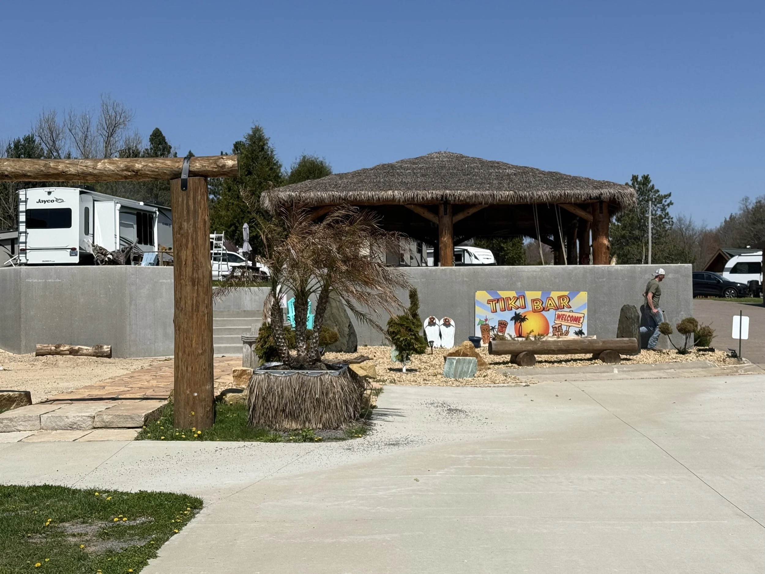 A tropical-themed outdoor tiki bar with a thatched roof, surrounded by trees and RVs, with a man walking in the vicinity and a colorful sign welcoming guests.