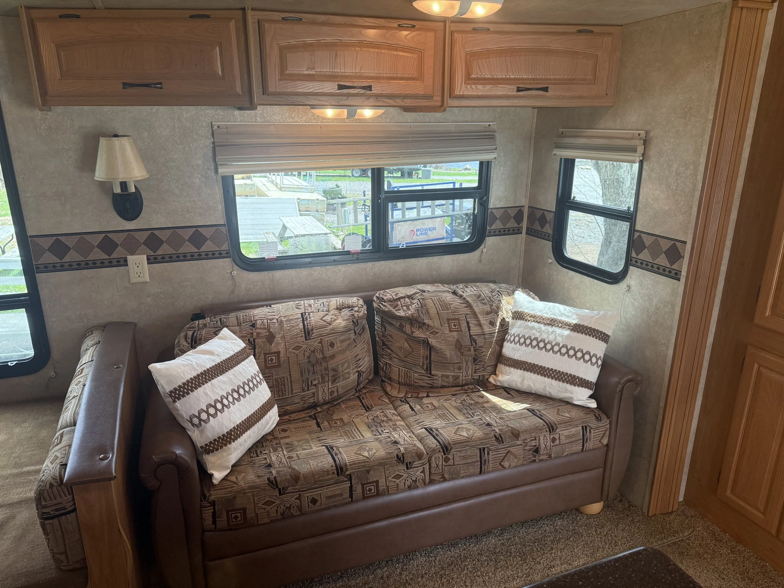 Interior of an RV or camper with a couch, two windows with blinds, wooden cabinets, and a wall-mounted lamp.