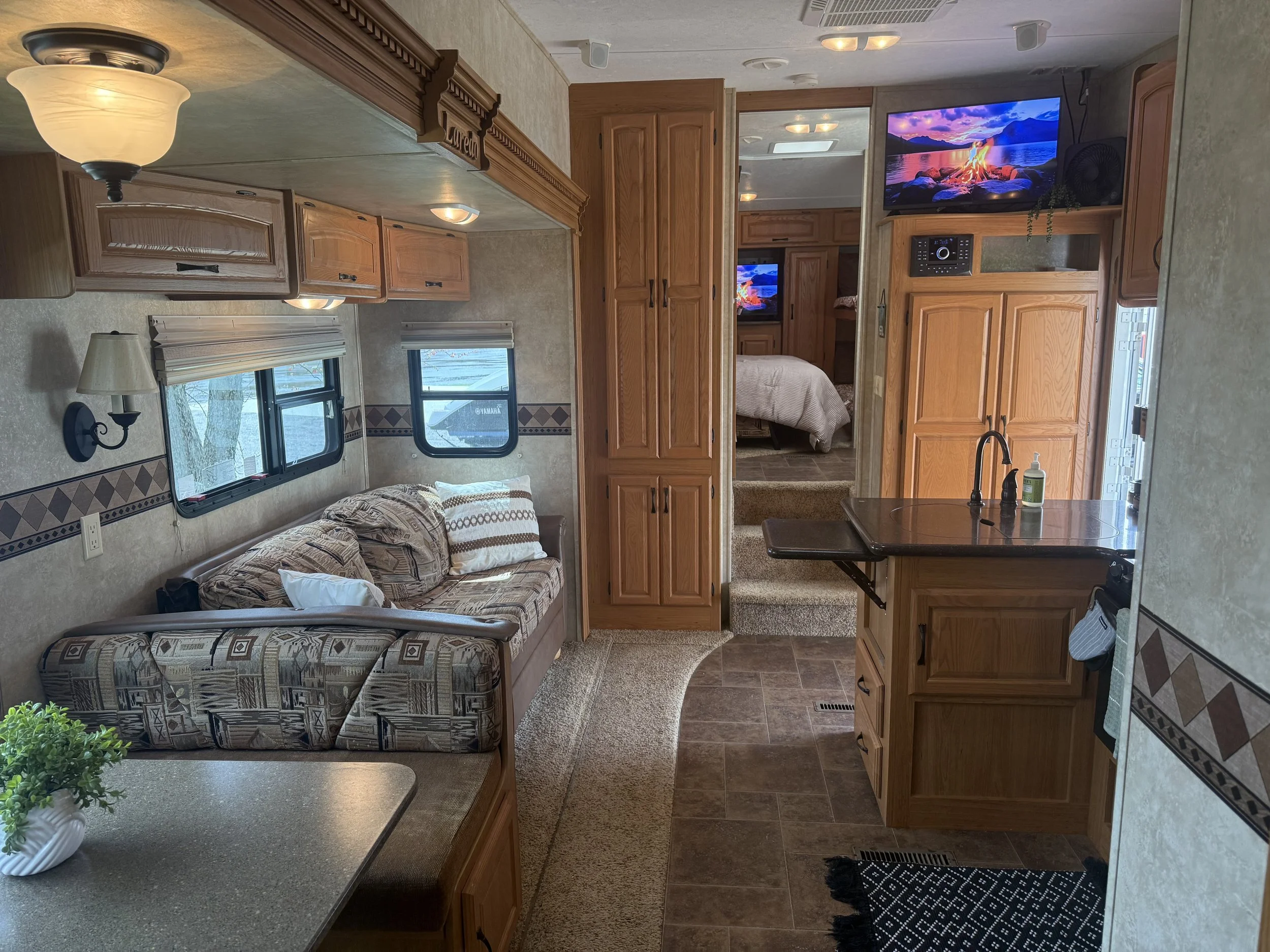 The interior of a cozy camper with wooden cabinets, a sitting area with a couch, a small table with a potted plant, and a kitchen counter with a sink and soap dispenser. A bedroom with a bed and TV is visible through an open door.