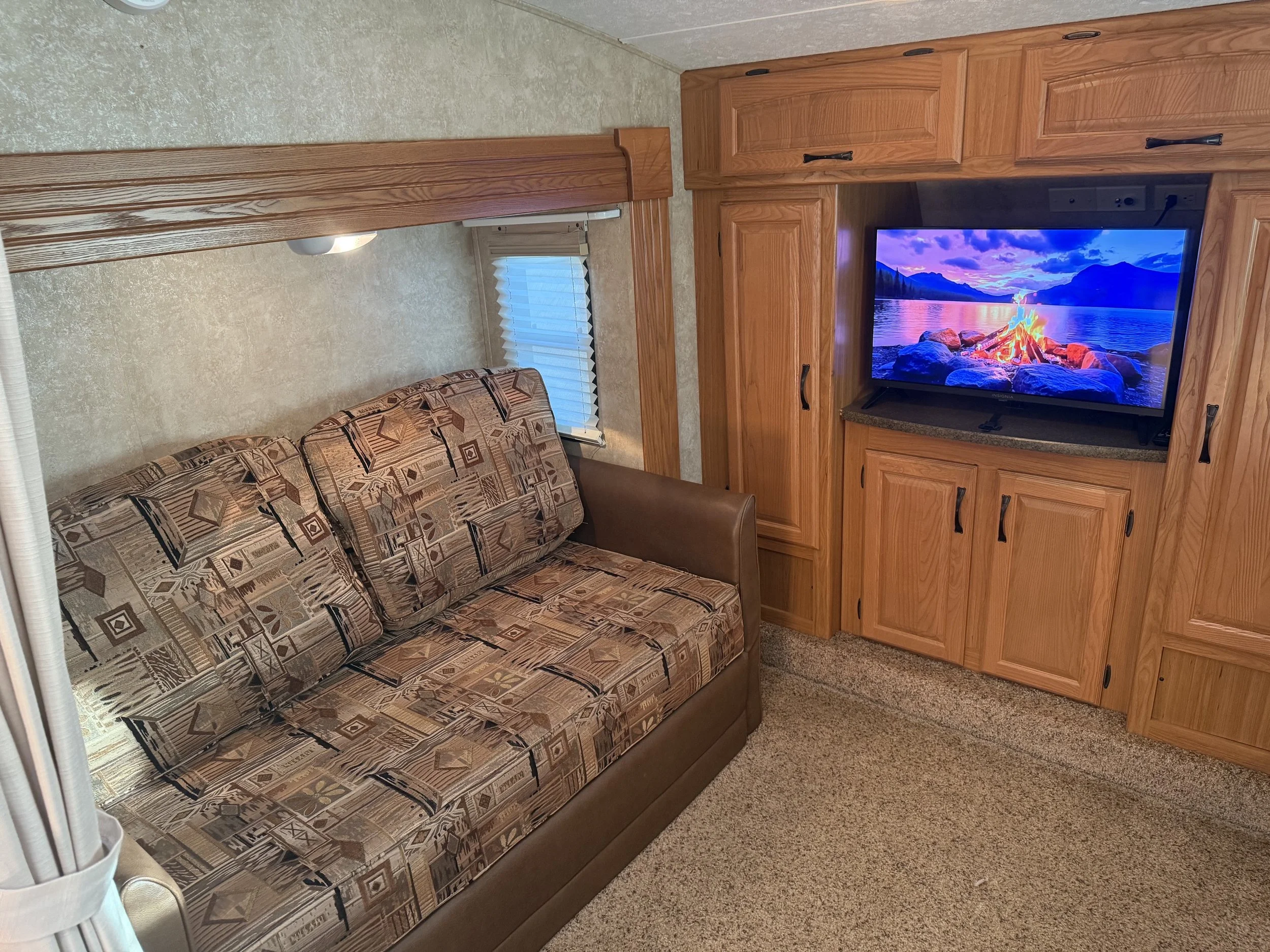 Living room area with a patterned sofa, a wooden entertainment center with a TV displaying a mountain and lake scene, a window with blinds, and beige carpet flooring.