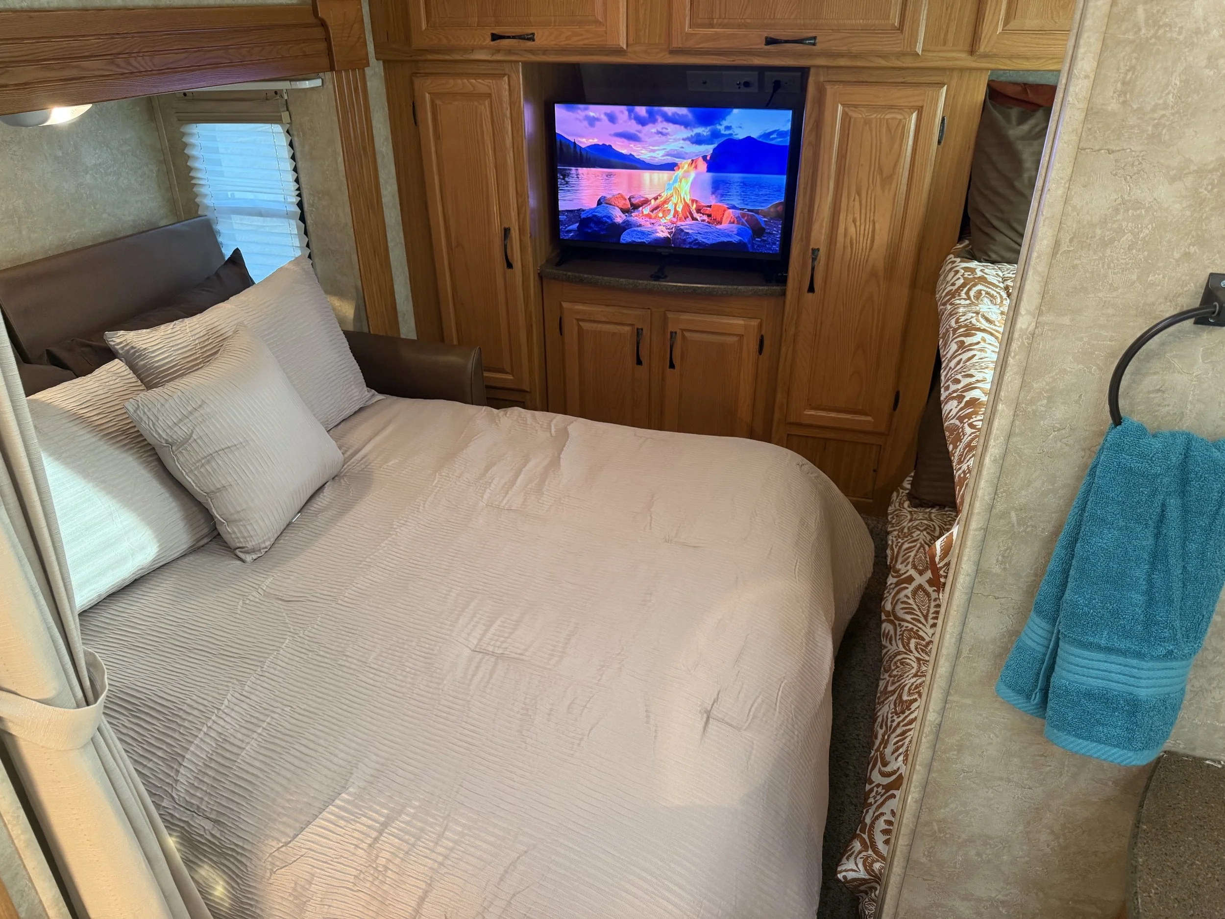 Queen-sized bed with beige striped bedding and multiple pillows, window with closed blinds, wooden cabinets surrounding a flat-screen TV displaying a scenic nature image, small seating area with patterned cushions, and washing supplies hanging on the