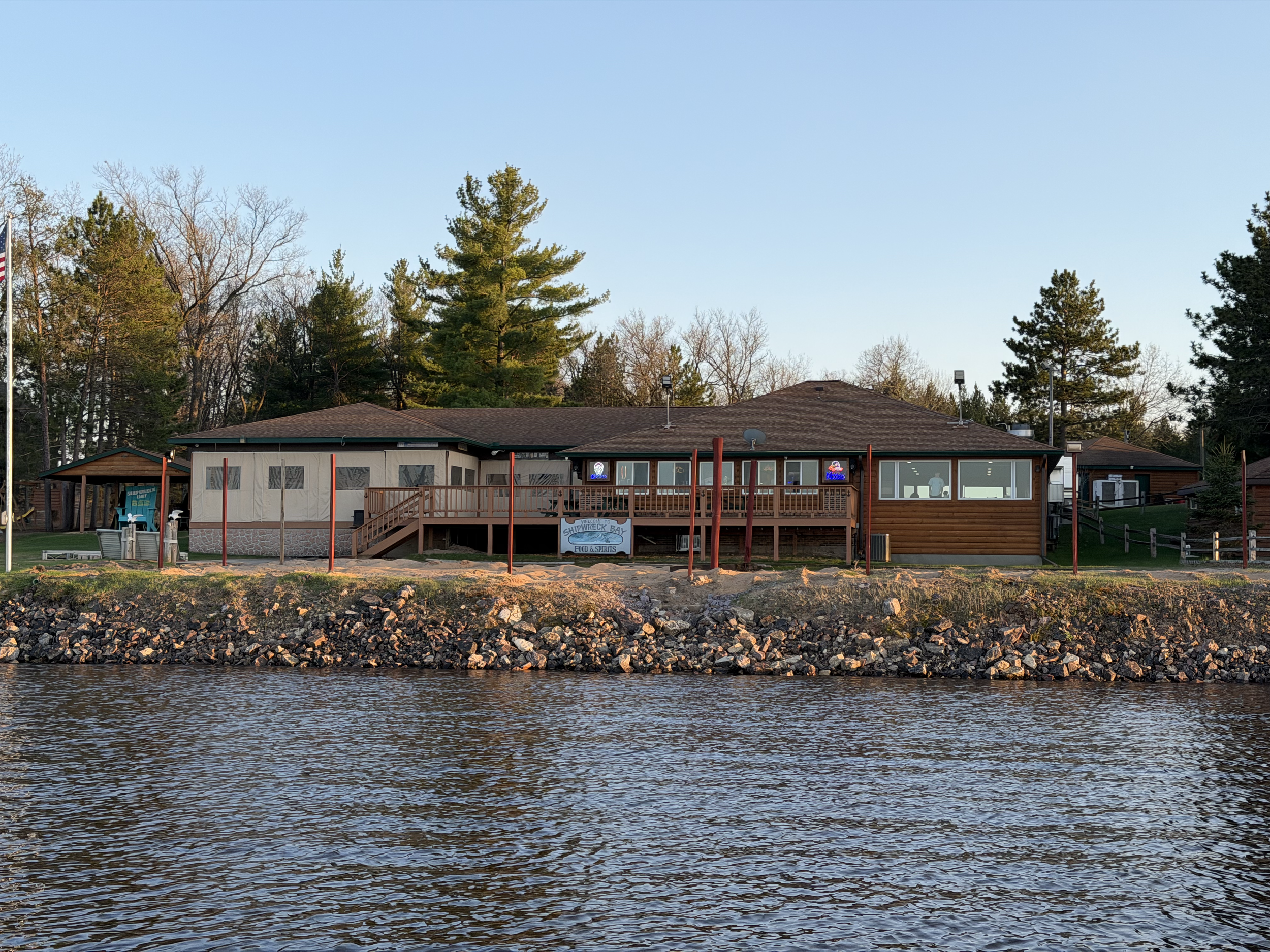 Waterfront view of a building with a wooden deck, trees in the background, and a sign for Shipwreck Bay Food & Spirits at sunset.