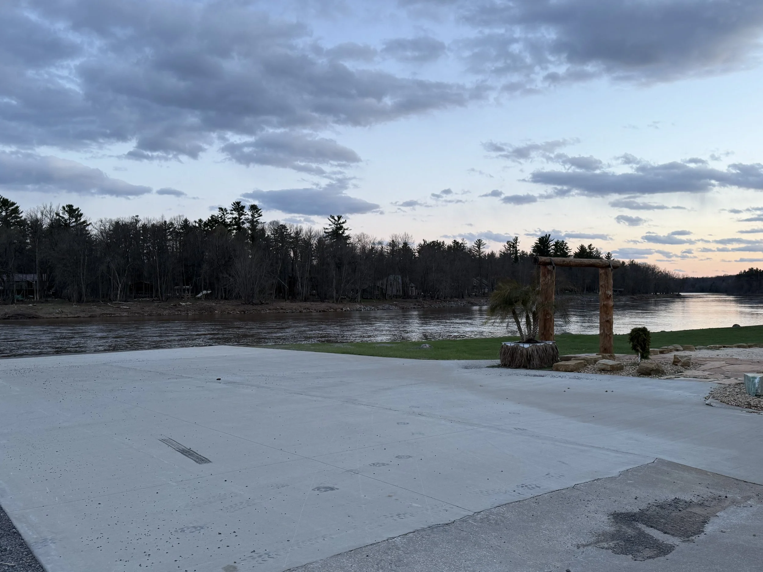 A scene by a river with cloudy sky, trees in the background, and a paved area in the foreground with tire marks. There is also a wooden structure on the right side and some small plants.