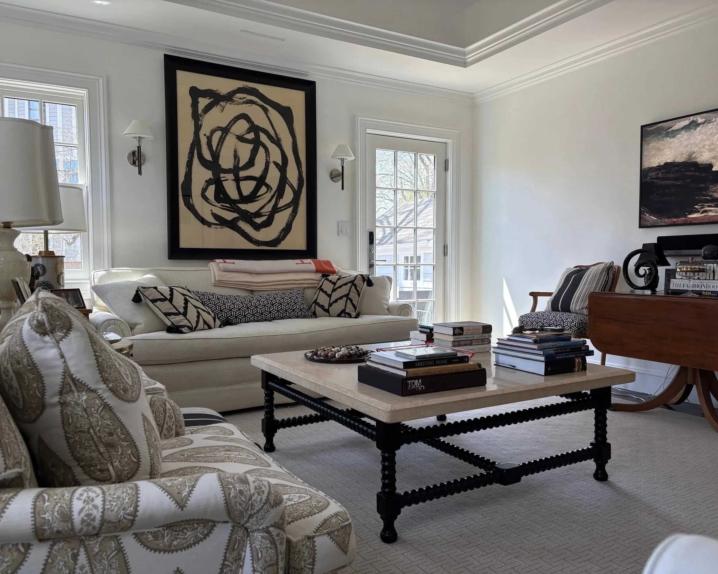 Using all neutral colors in living room to rest your eyes and focus on Frame tv art and entertainment, large windows, a sofa with patterned pillows, a marble coffee table with books and decorative items, and abstract artwork on the walls.