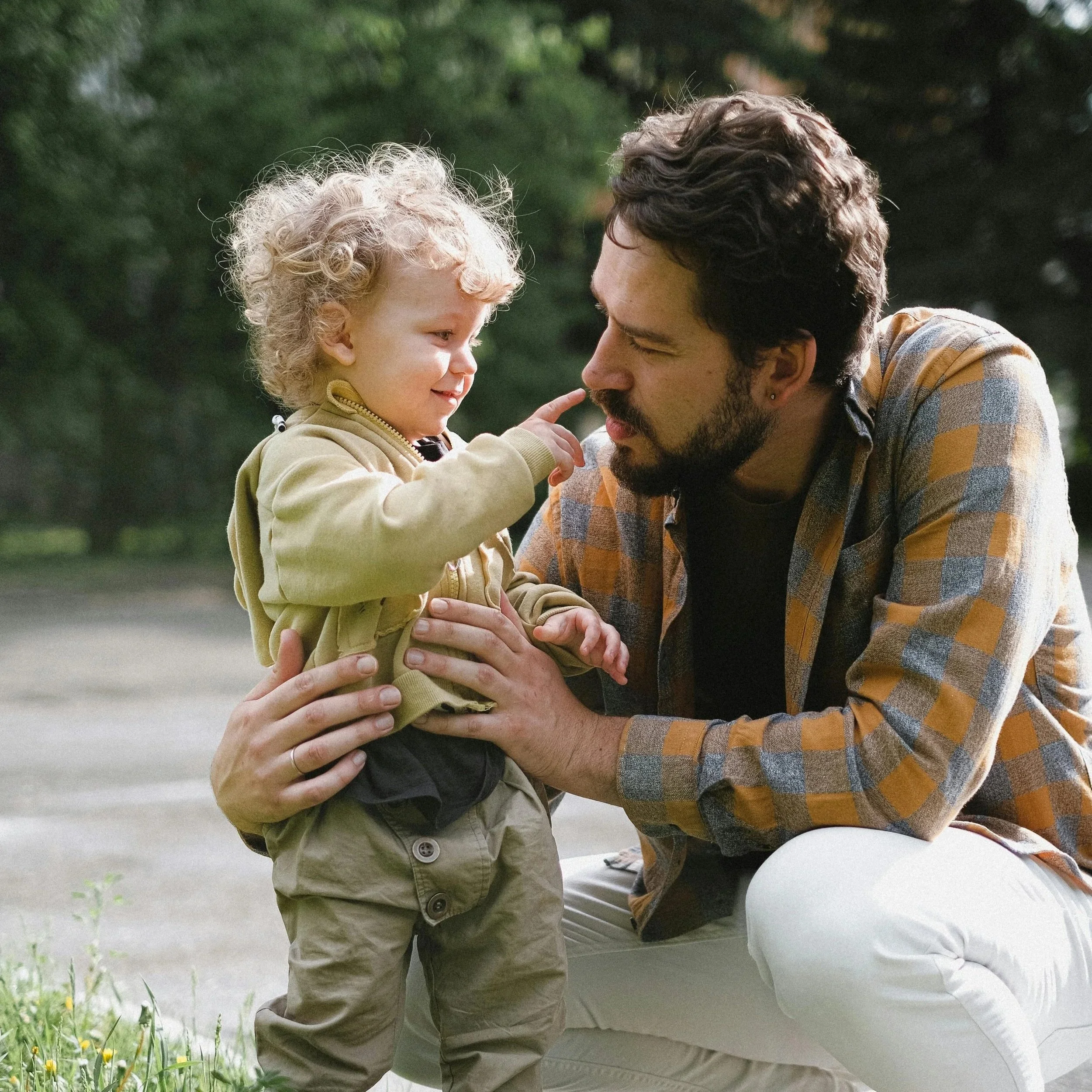 A young child with an adult outdoors near a green leafy background. The child is touching the adult's face, and they are engaged in a tender moment.