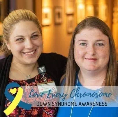 Jessica Duis and another woman smiling at the camera at a Down syndrome awareness event. One has blond hair and the other has brown hair. There is a yellow awareness ribbon and a blue banner that says 'Love Every Chromosome'.