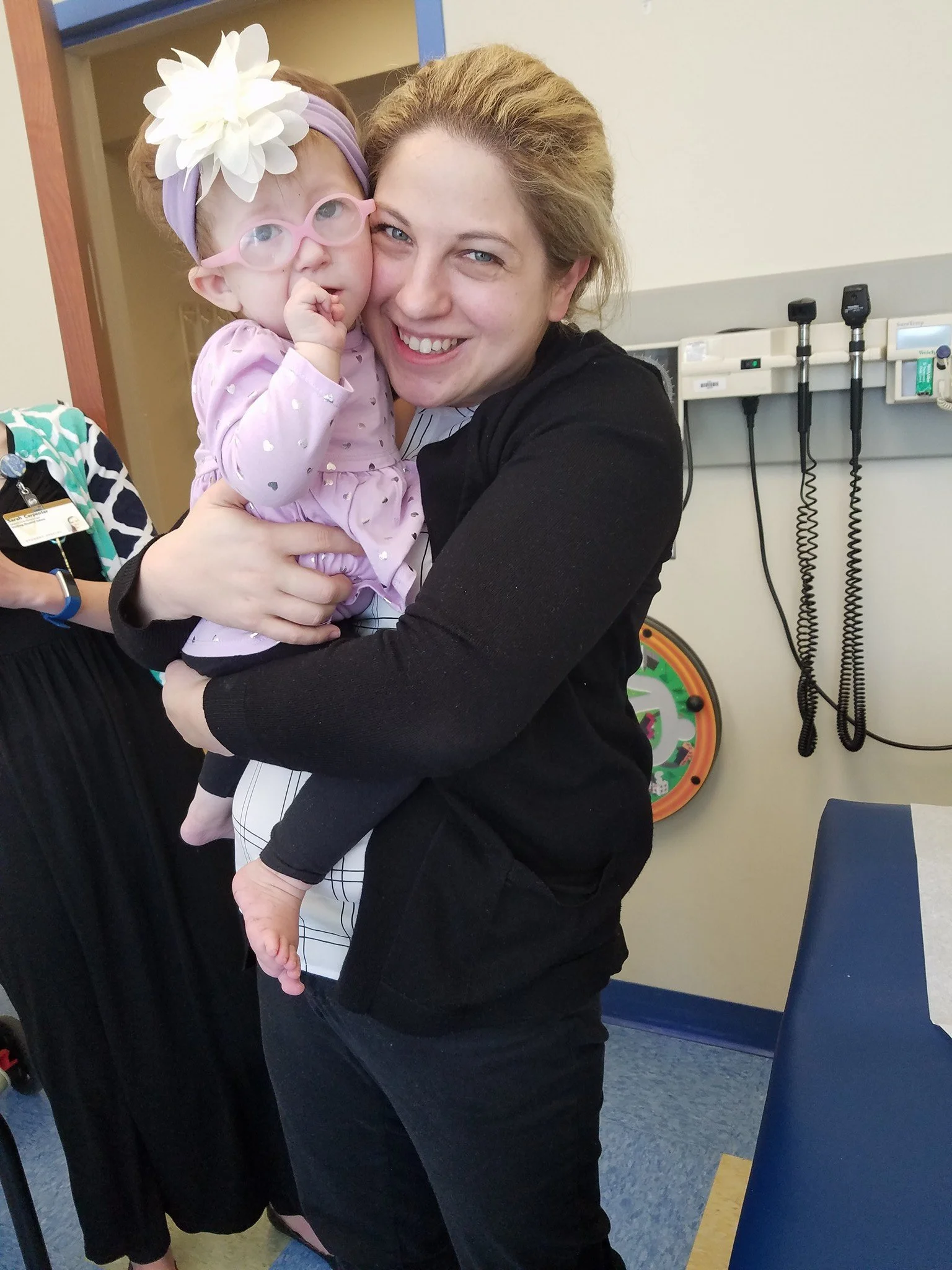 Dr. Jessica Duis is holding a little girl in a medical setting, with medical equipment on the wall behind them.