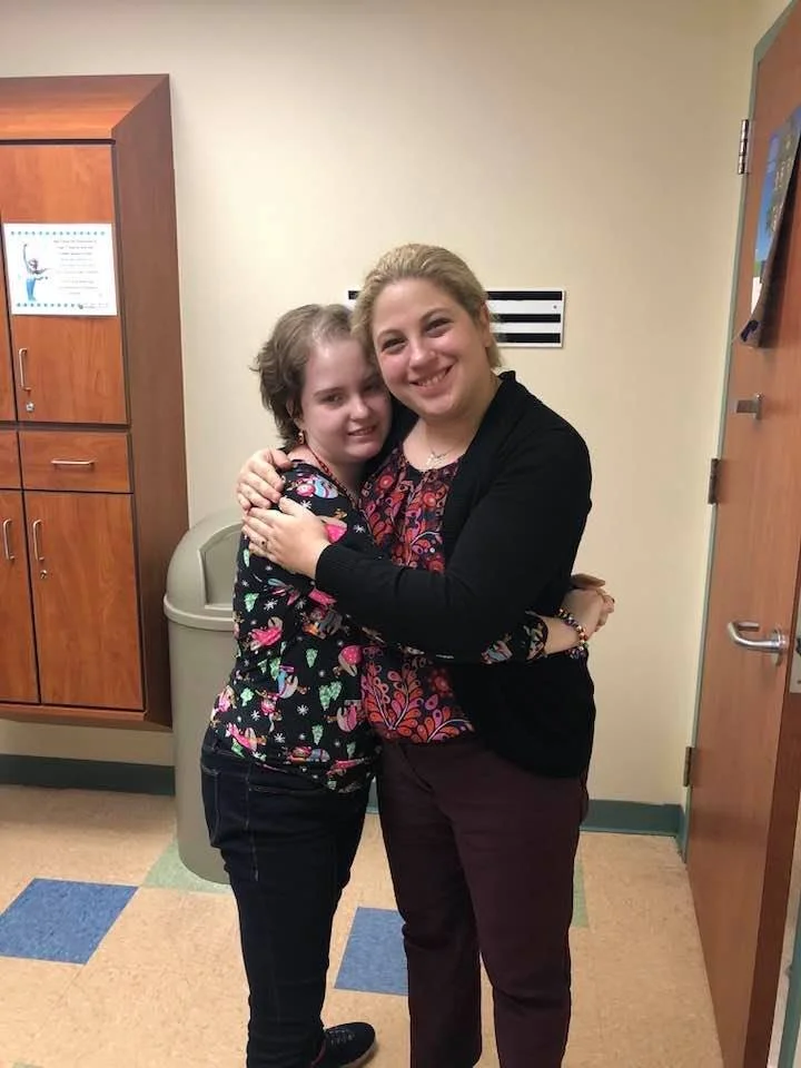 Jessica Duis hugging a patient and smiling in an indoor setting, possibly hallway or room, with cabinets and a trash can behind them.