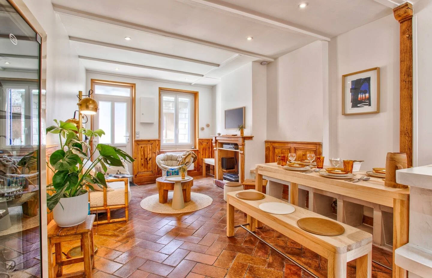 Bright living room with white walls, wooden accents, a fireplace, a wall-mounted TV, a large window, and a sitting area with a chair and small table. There is also a dining area with a wooden table, benches, and tableware, plus a houseplant.