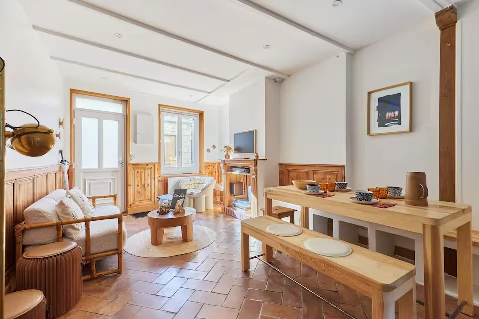 Bright living room with white walls, wooden accents, a small sofa with cushions, a round coffee table, a fireplace, a TV, and a dining area with a wooden table and benches, decorated with bowls and a pitcher.