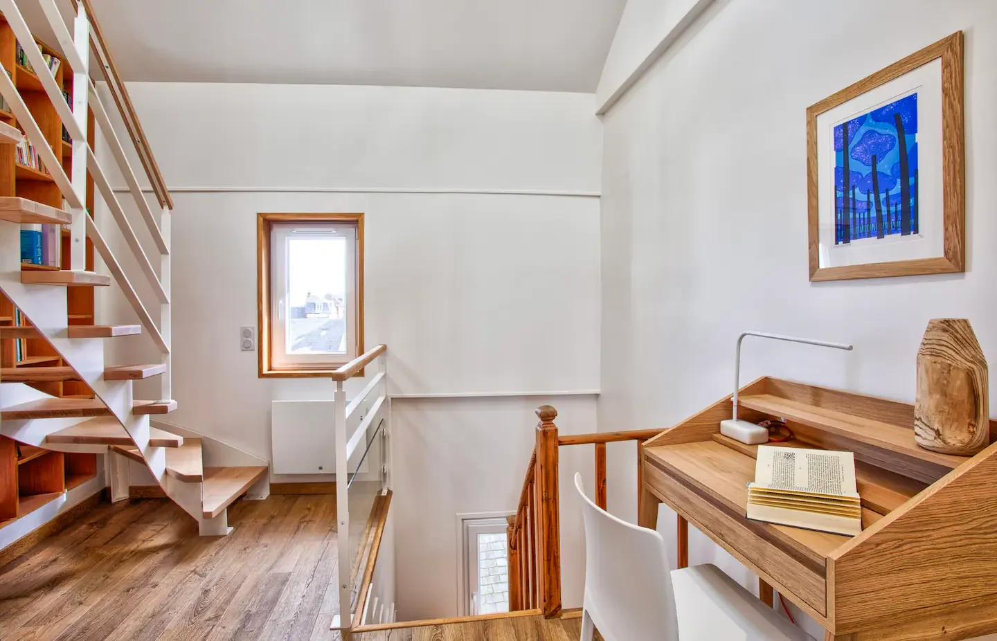 A cozy home corner with a wooden desk, white chair, and a lamp, decorated with a wooden vase and an open book, near a staircase and window with a city view.