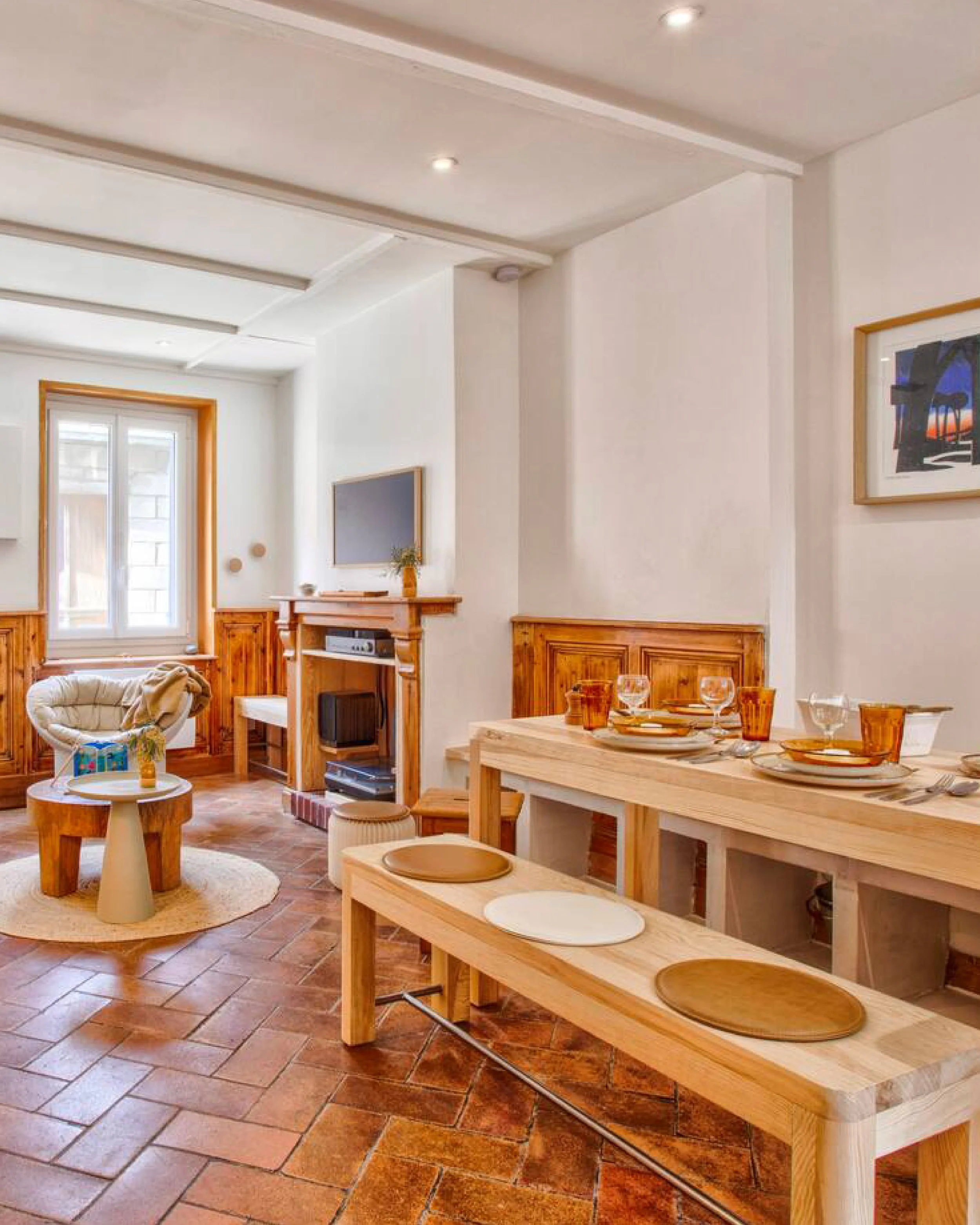 A cozy living room with wood-paneled walls, a flat-screen TV above a fireplace, a window with wooden trim, a small round table with decorative items, and a wooden dining table set for meals. The room features warm tones and tiled flooring.