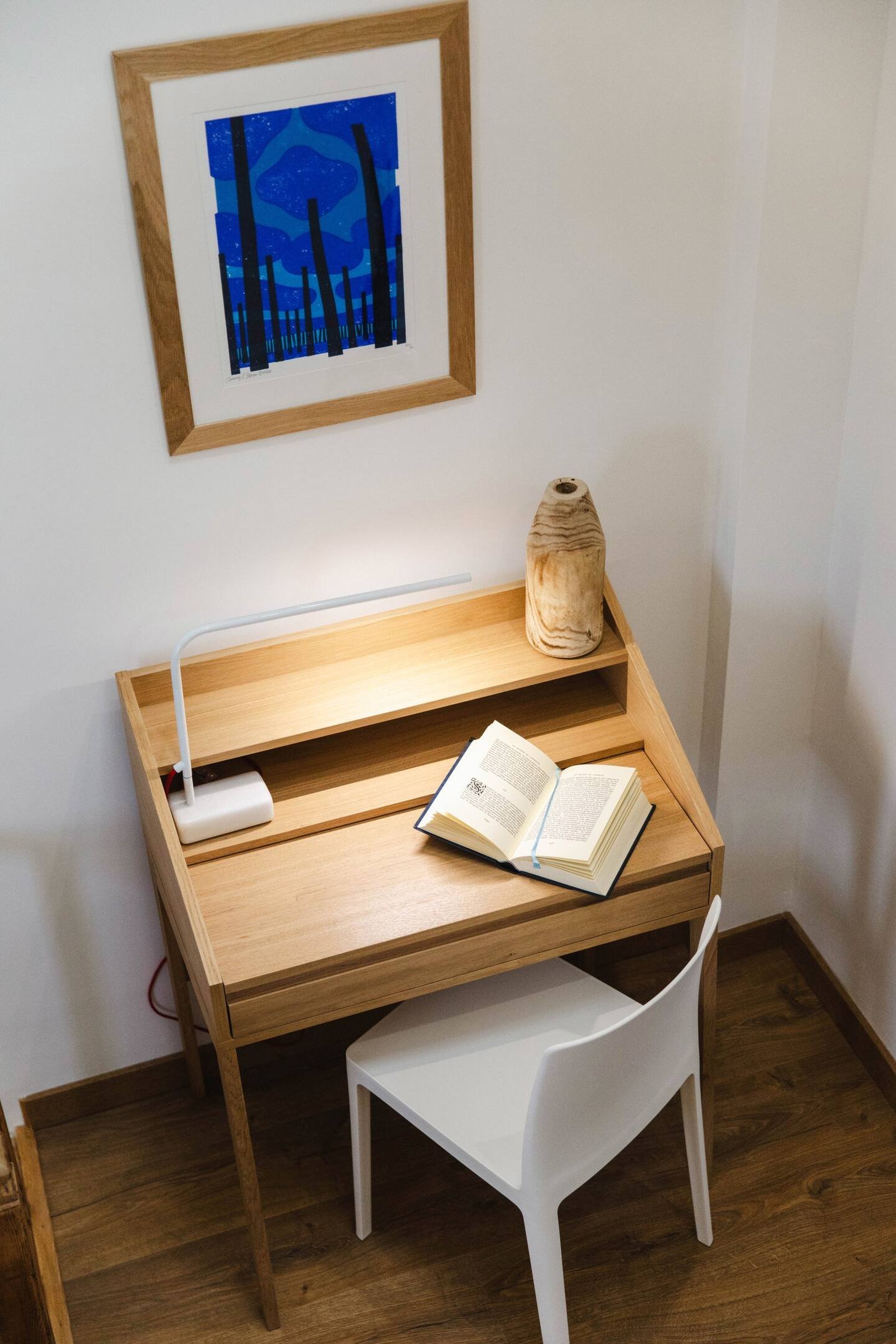A small wooden desk with a book open on top, a white chair, a table lamp with a white base, a wooden vase, a framed abstract artwork in shades of blue on the wall, and a corner of a wall with white paint and wooden flooring.