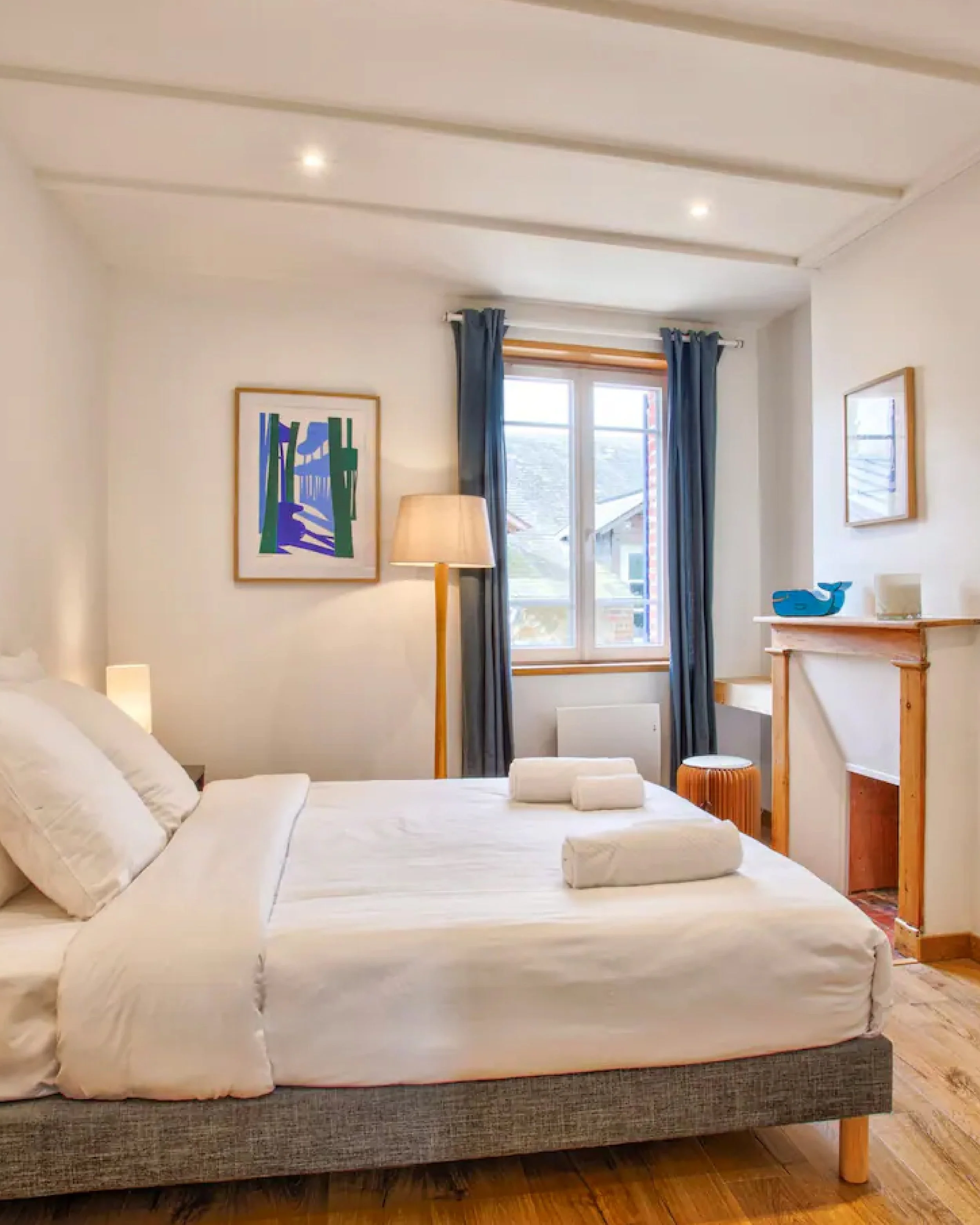 A bedroom with a large bed covered in white linens and pillows. There is a window with blue curtains and artwork on the walls. A floor lamp, a wooden table with a blue whale figurine, and a small stool are also visible.