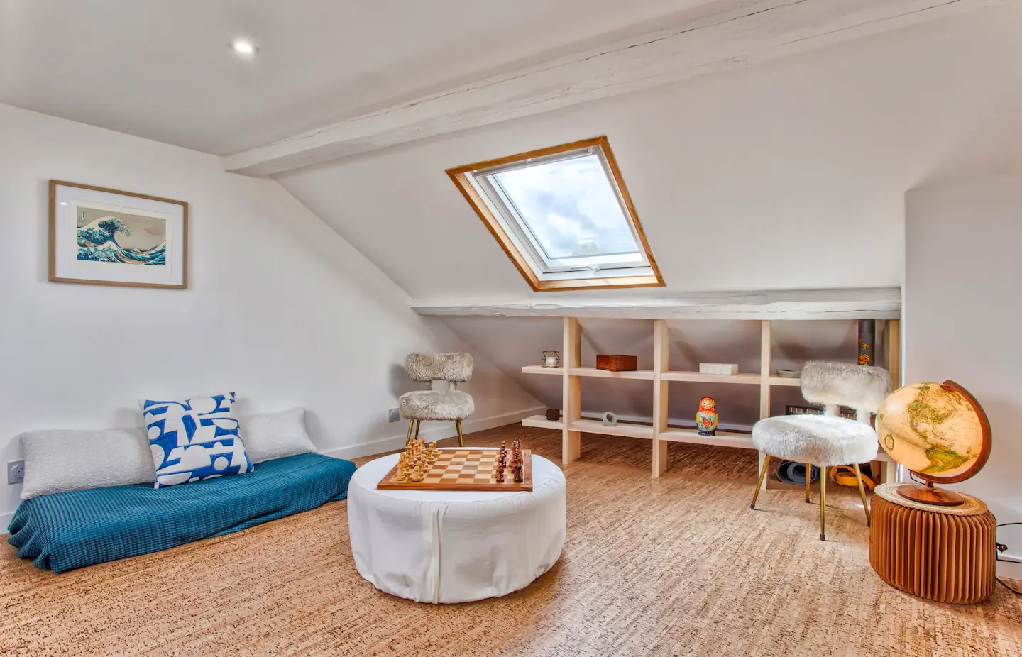 Attic room with a skylight, white walls, and a beige carpet. Features a low white couch with blue and white cushions, a round table with a chessboard, two furry chairs, a globe on a wooden stand, a built-in shelf with decorative items, and a framed p