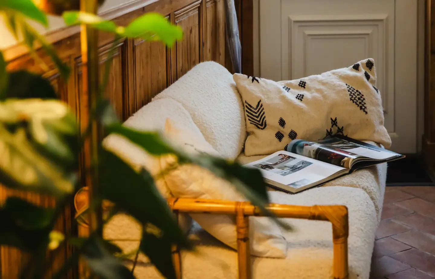 A cozy living room with a beige sofa featuring black and white decorative pillows, an open magazine on it, a terracotta tiled floor, and wood-paneled walls. Some green leaves are partially visible in the foreground.