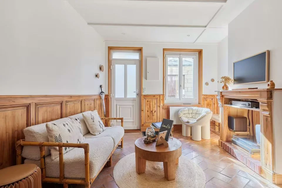 Cozy living room with wooden paneling, a white sofa with decorative pillows, a small round wooden coffee table, a cushioned chair, a fireplace, a wall-mounted TV, and large windows.