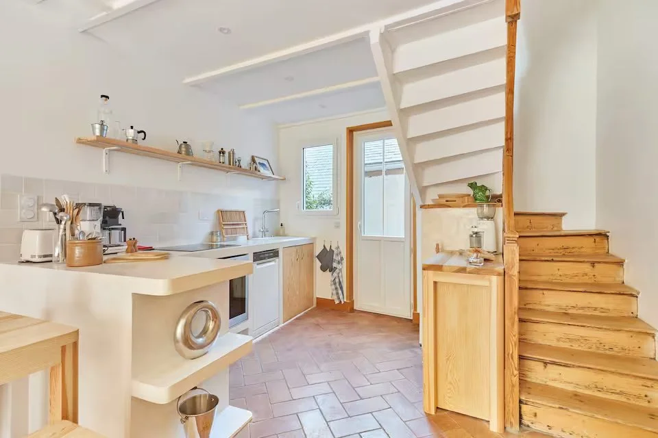 Kitchen with white walls, wooden cabinets, and a staircase leading upstairs. There are open shelves with kitchenware, a window and door leading outside, and a countertop with appliances and utensils.