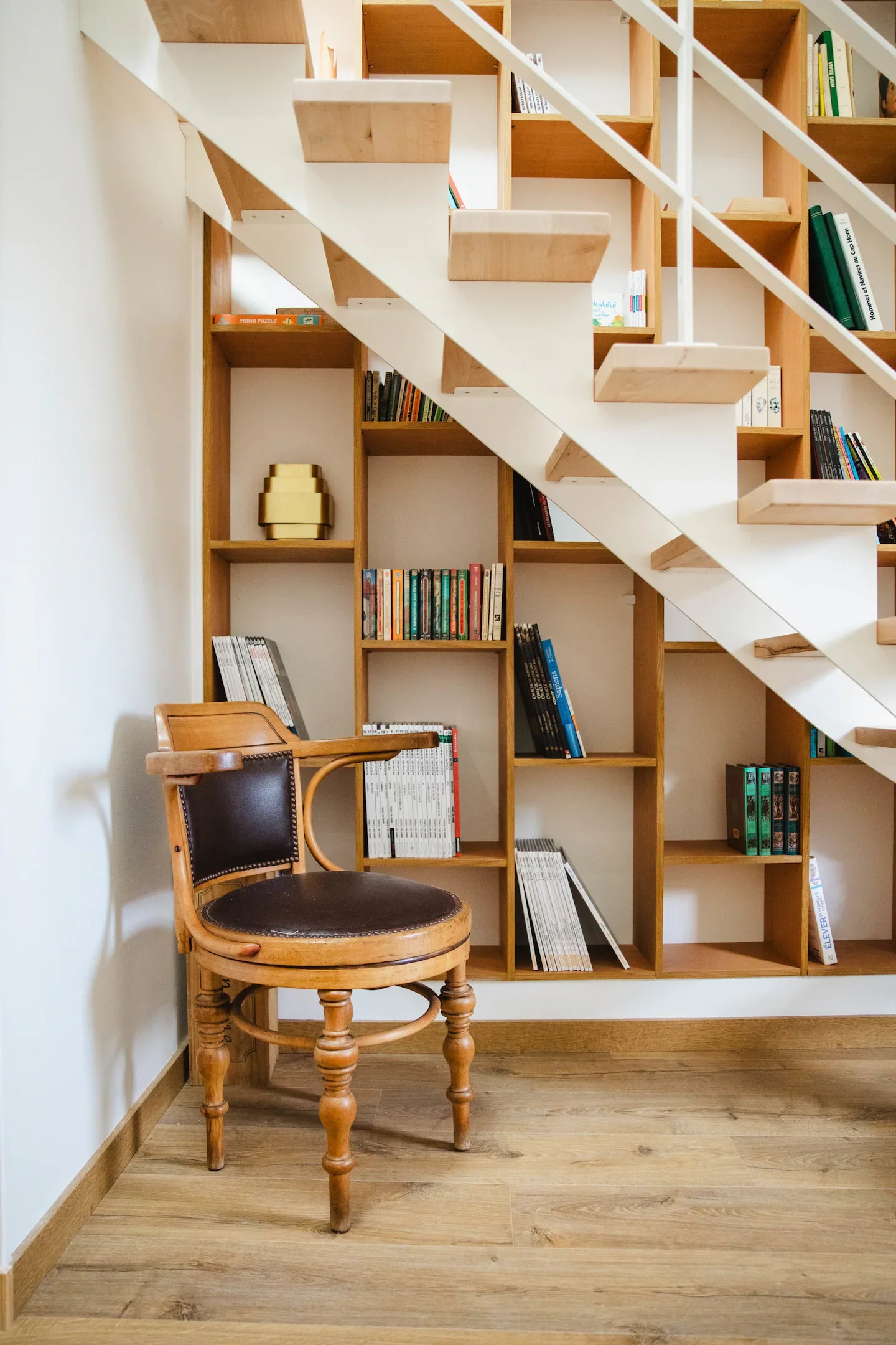 A wooden bookshelf filled with books and magazines behind a vintage wooden chair with a leather seat and backrest, in a room with wooden flooring and white walls.