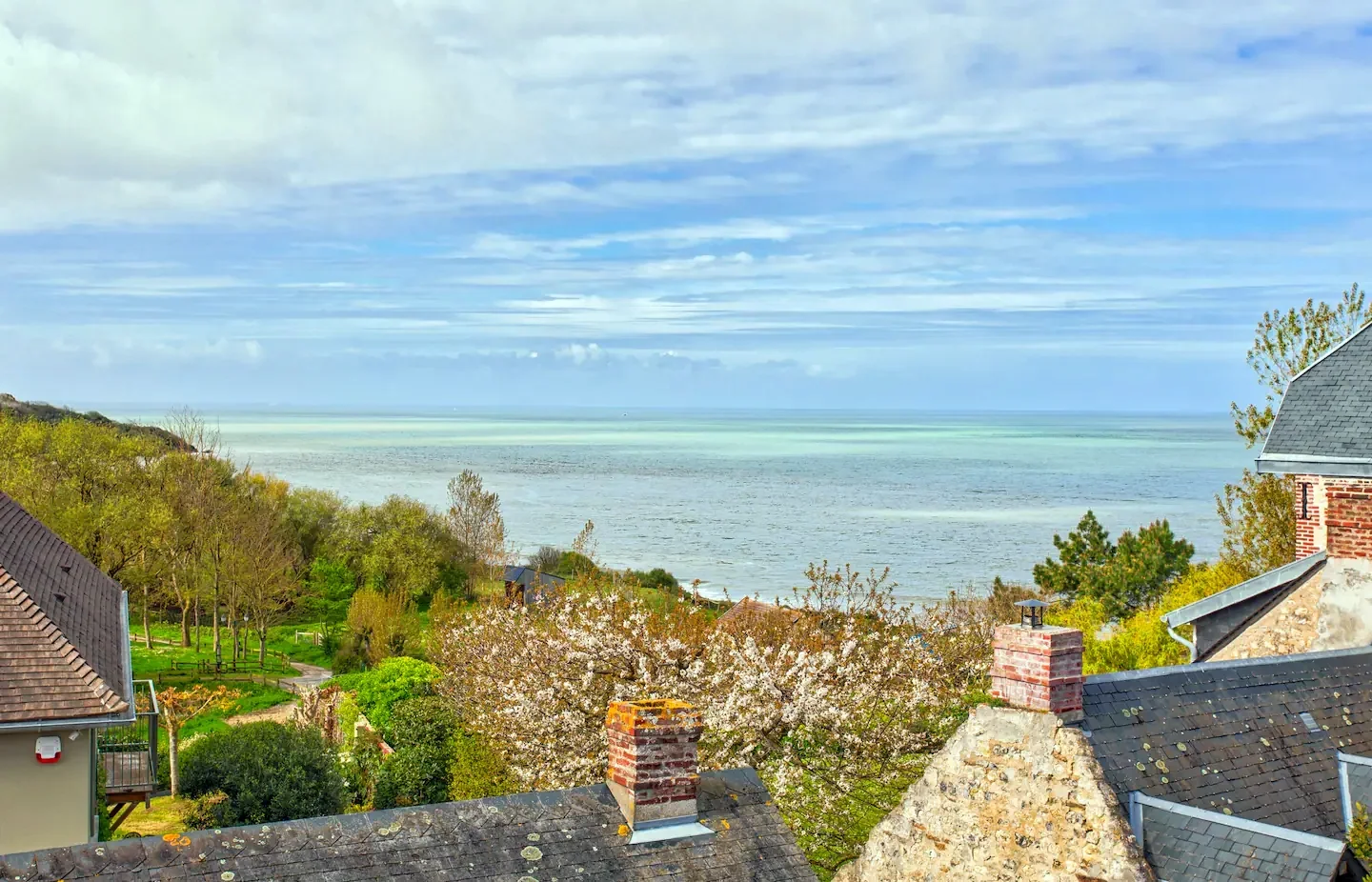 A view of a coastal landscape with houses, trees, and the sea in the distance, under a partly cloudy sky.