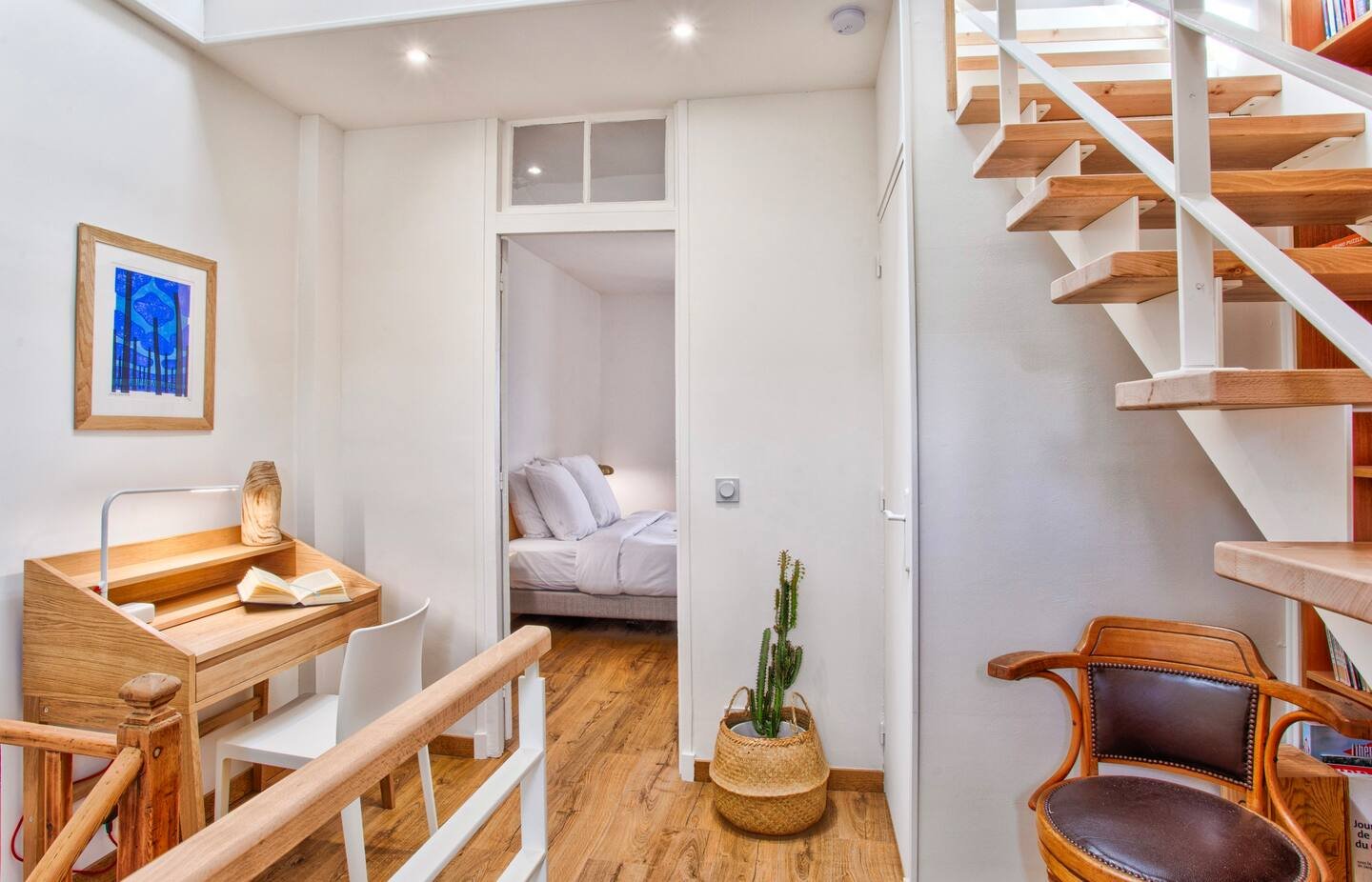 Interior view of a small apartment with a bedroom visible in the background, wooden staircase on the right, a small wooden desk with a white chair on the left, a cactus in a basket in the center, and a wooden armchair near the stairs. The space featu