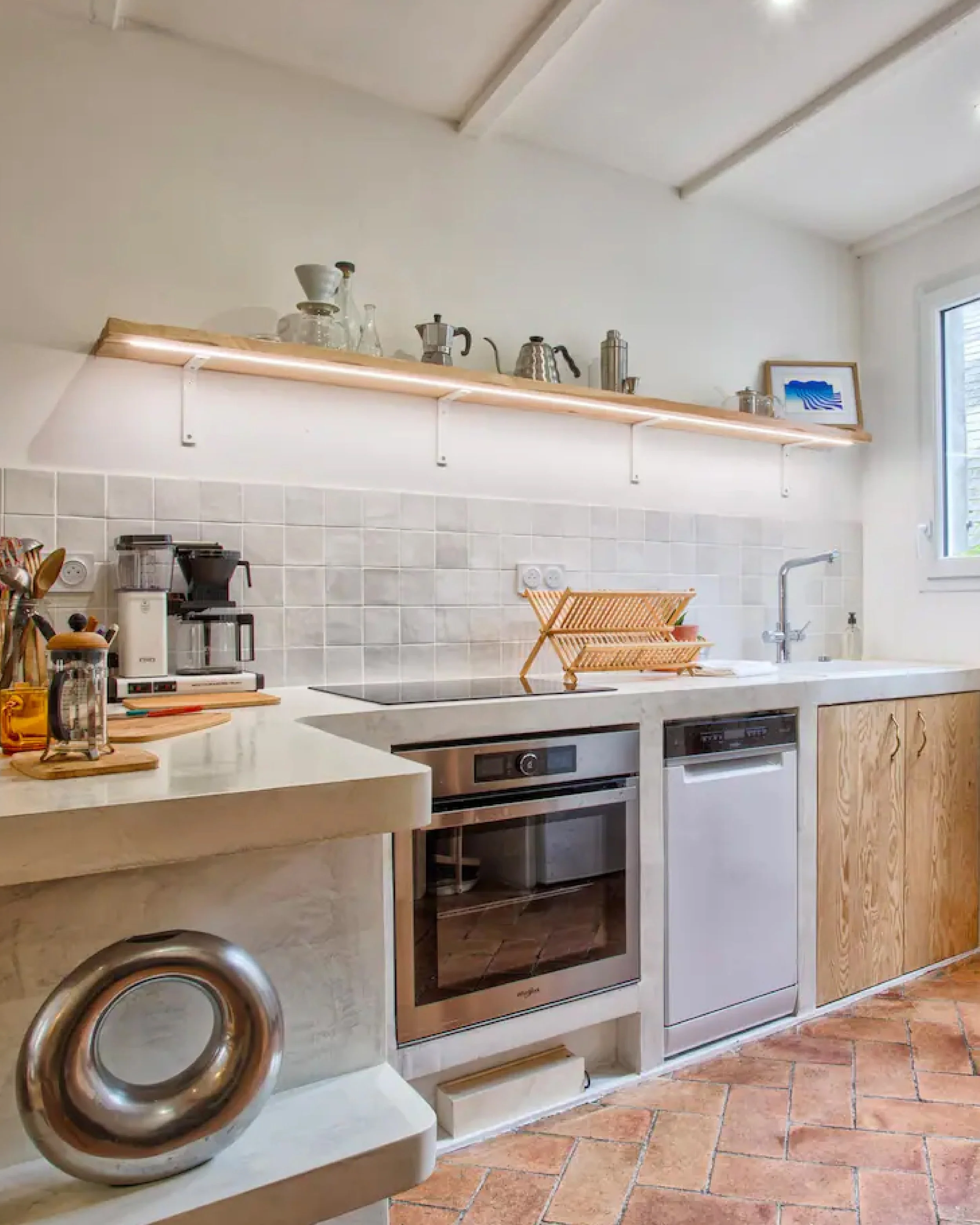 A kitchen with a beige countertop, a built-in oven, white tiled backsplash, and a wooden shelf on the wall holding various coffee pots and a picture frame. There is a window on the right side and a copper-colored object in the foreground.