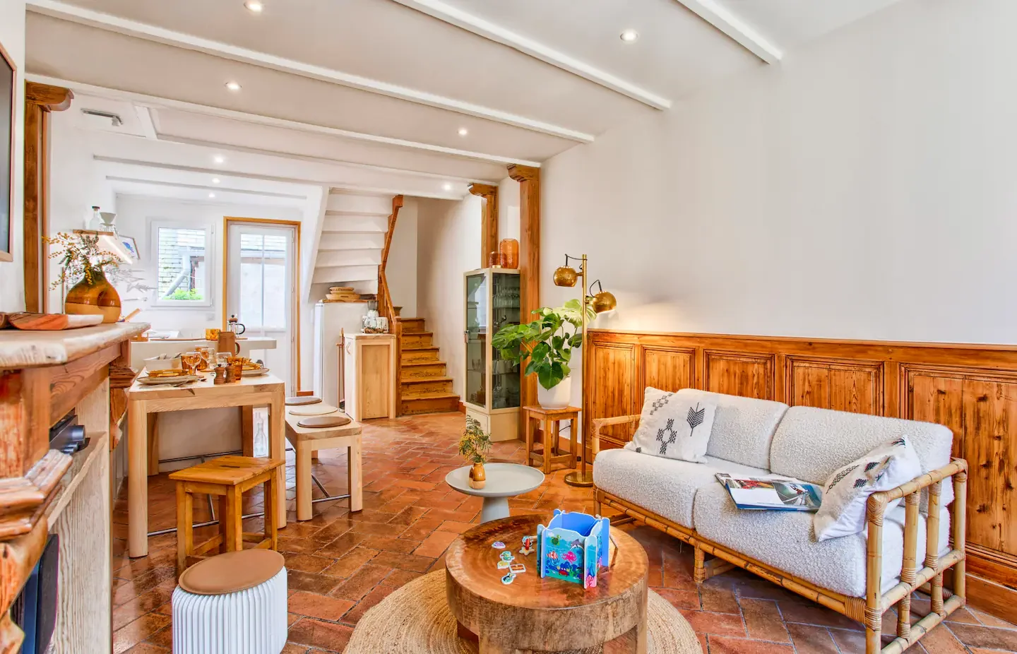 Bright living room with wooden accents, white walls, and terracotta tiled floor, featuring a sofa, wooden dining table, and staircase.
