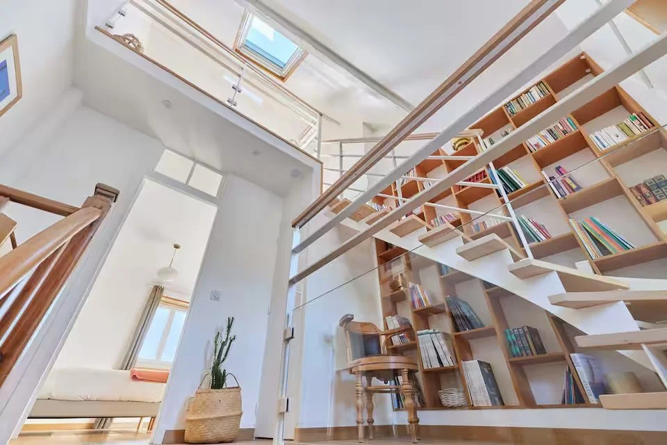 Interior view of a modern home with a curved wooden staircase, a built-in bookshelf filled with books, a small wooden desk, and a high ceiling with skylights.