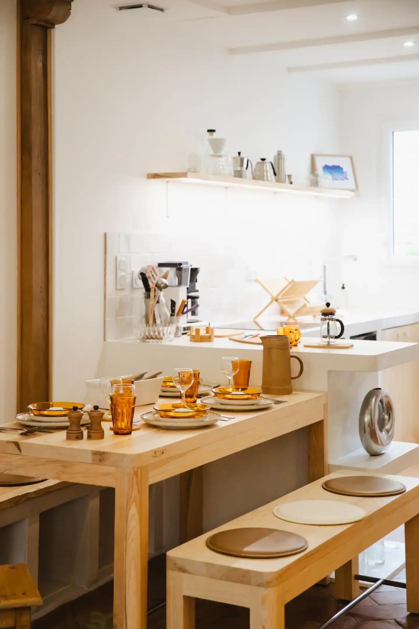 Kitchen area with a wooden table set for a meal, including plates, glasses, and utensils. The background features a white wall with a shelf holding various kitchen items and a framed picture. There are wooden chairs or benches with round seats and a 