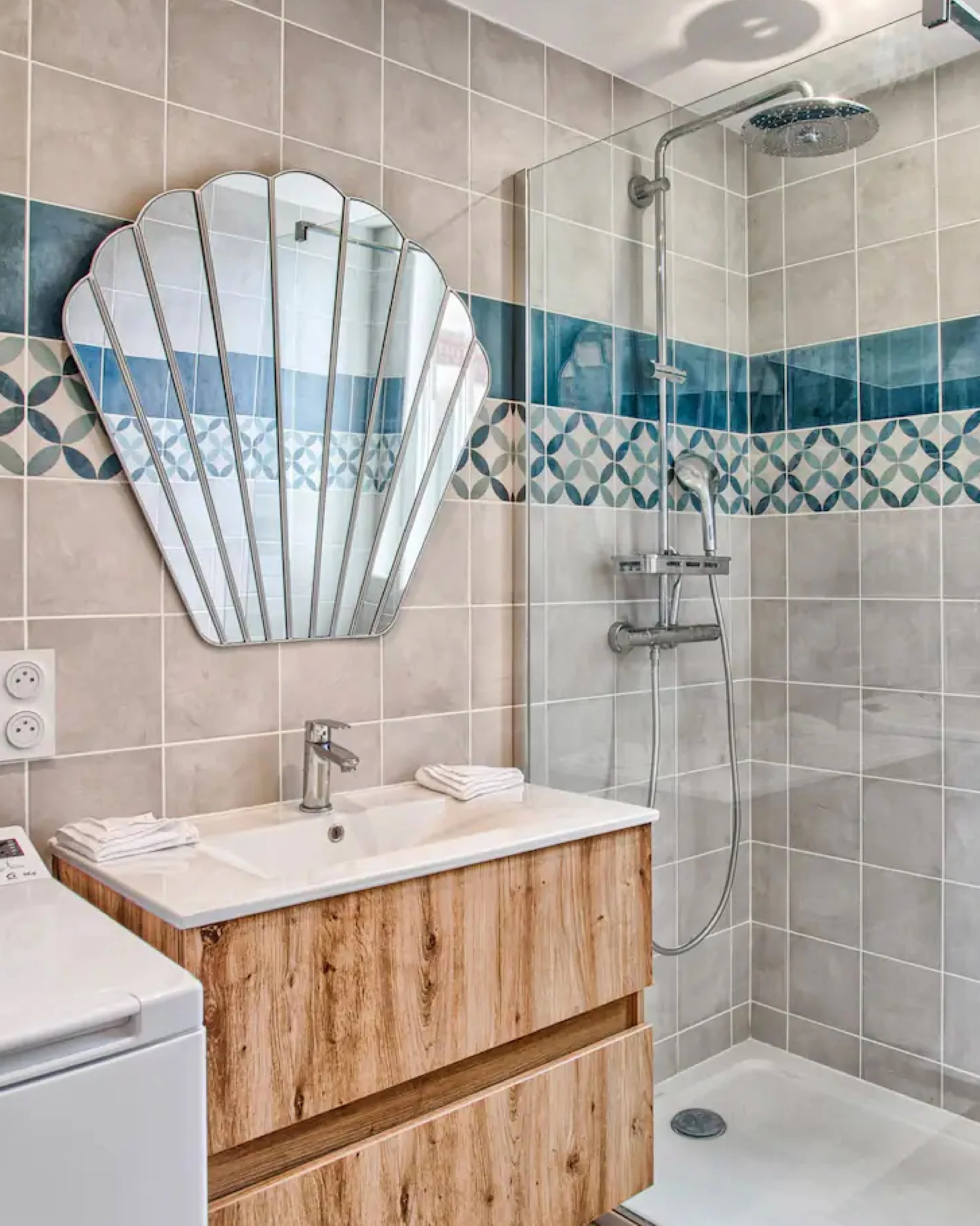 Bathroom with light-colored tiled walls accented by a horizontal strip of decorative tiles, a vanity with a wooden finish and a rectangular sink, a mirror with a scalloped edge, a shower area with a large round showerhead, a handheld shower, and a sm
