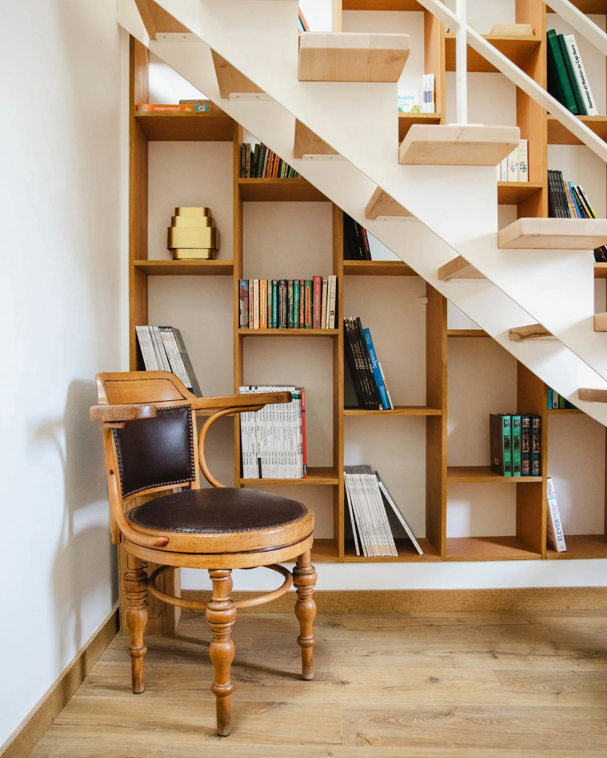 A wooden chair with a cushioned leather seat and backrest in front of a large wooden bookshelf filled with books, magazines, and three decorative gold bowls, with a light-colored wall and wooden floor.