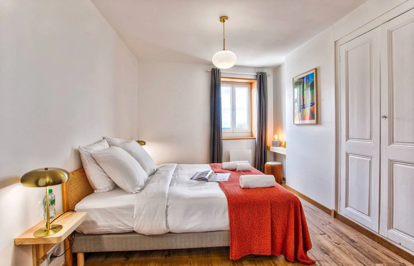 A cozy bedroom with a double bed, white bedding, and a red blanket, topped with neatly folded towels and an open magazine. Gold bedside lamps flank the bed. A window with gray curtains lets in natural light. A small desk with a lamp and decorative it