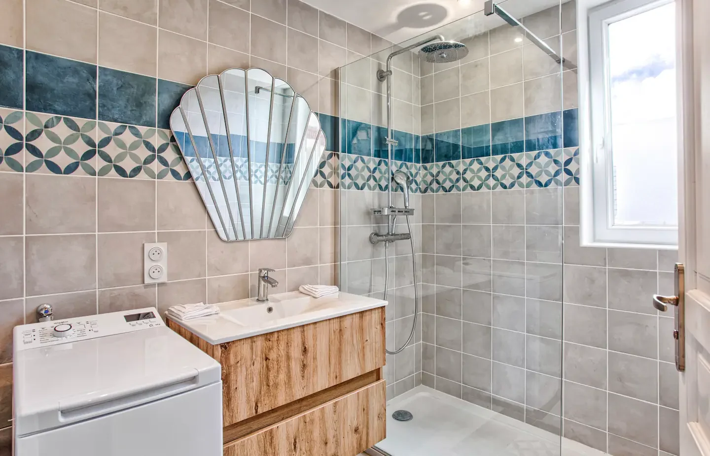 Bathroom with shower area, wall-mounted mirror, wooden vanity with double sinks, washing machine, and a window.