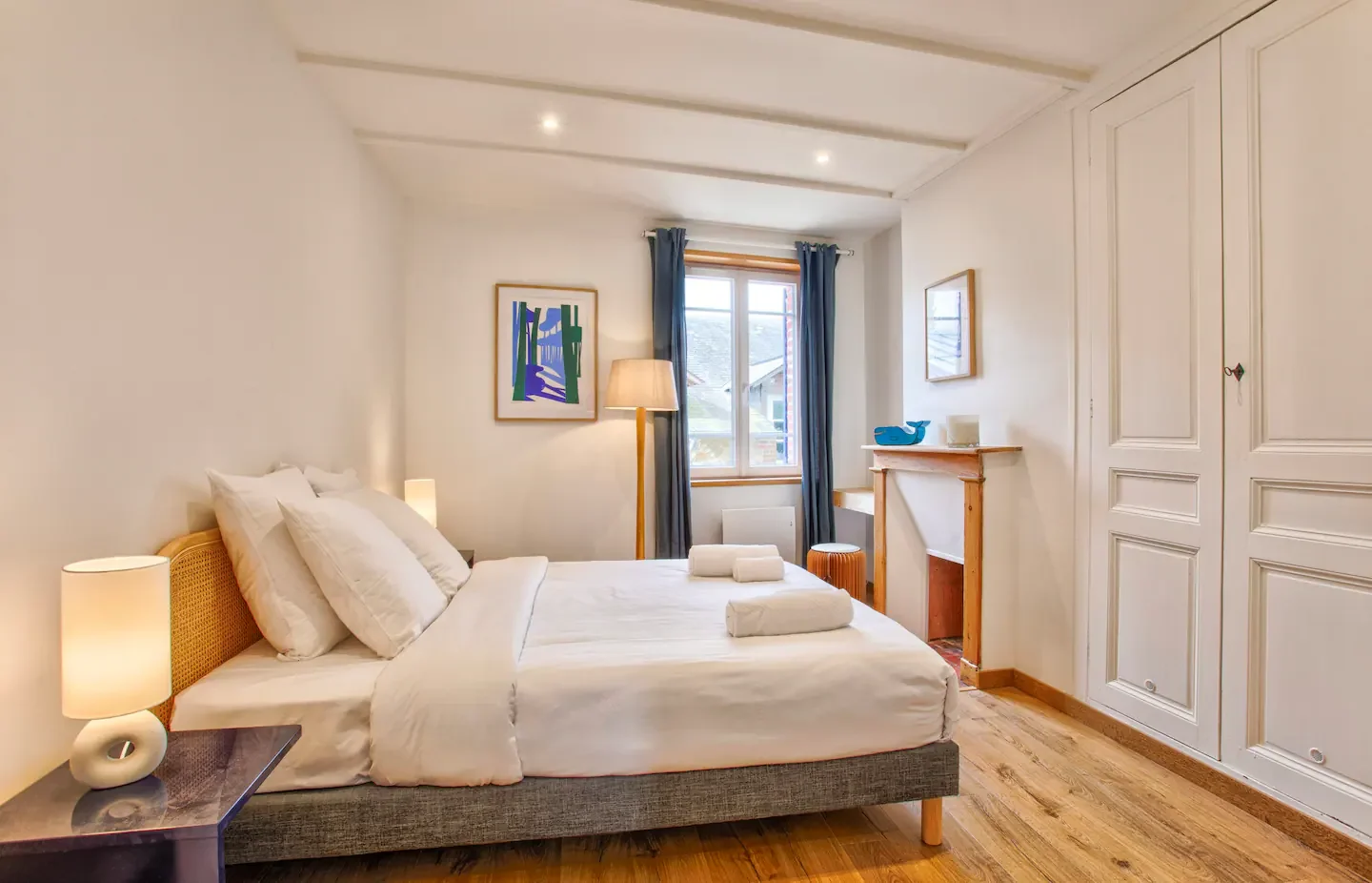 A cozy bedroom with a large bed, white bedding, and multiple pillows. Two nightstands with lamps, a window with blue curtains, and artwork on the walls. There is a wooden floor and built-in white closet doors.