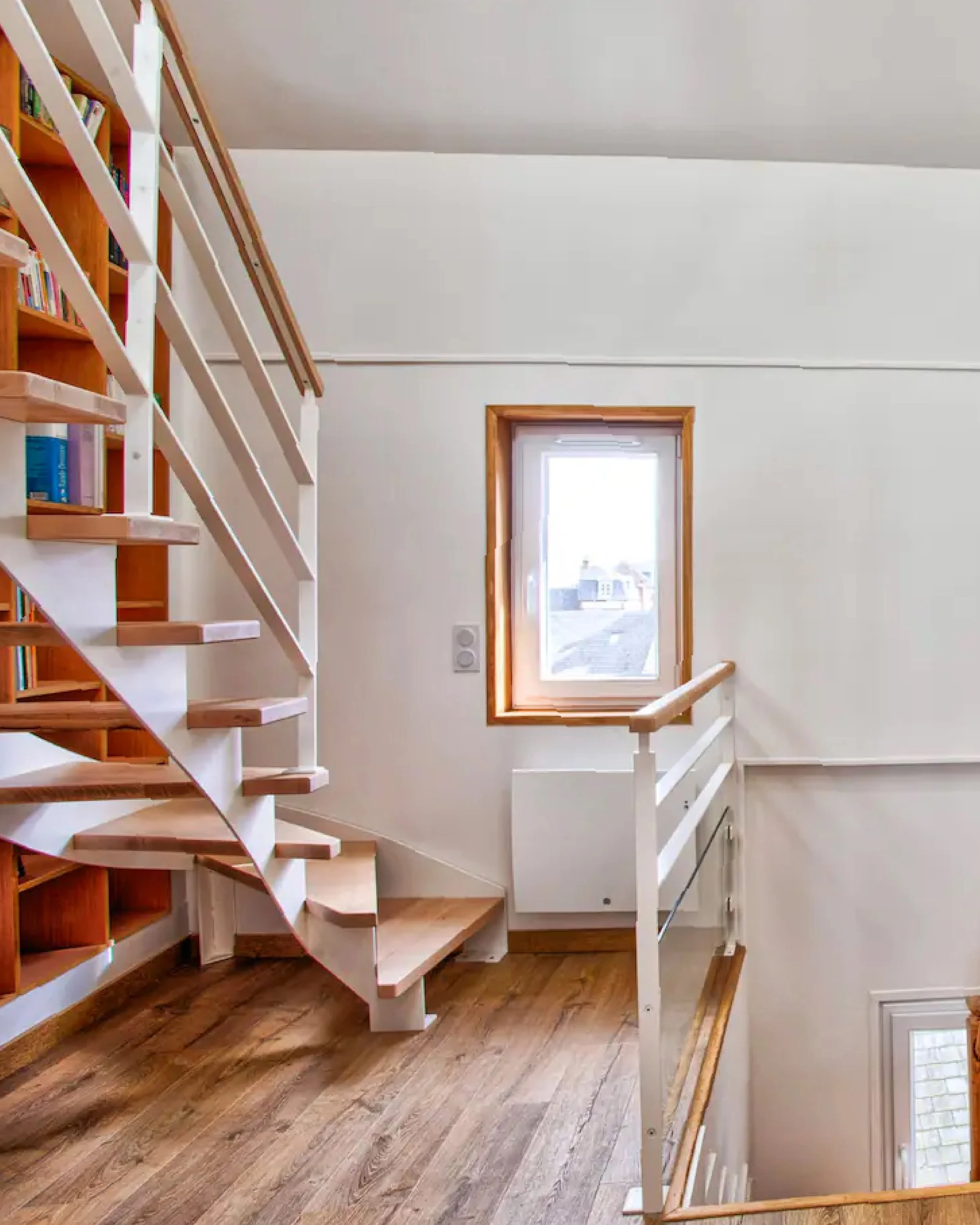 Interior of a house featuring a staircase with wooden steps and white rails, situated next to a wall with a window allowing natural light, and wooden flooring.