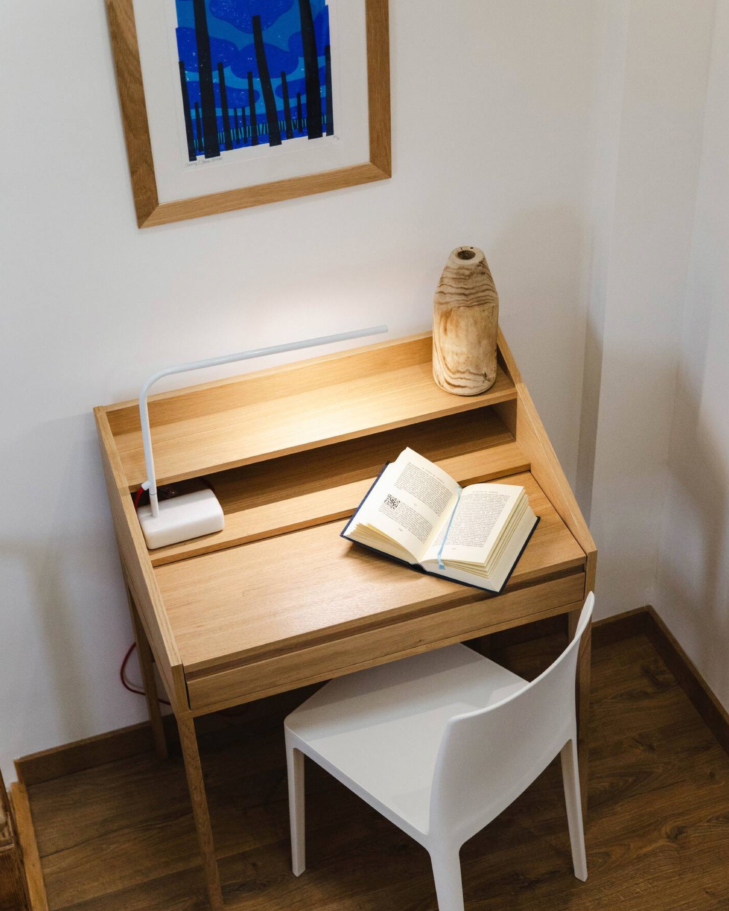 A wooden desk with a white chair in front and a black chair hidden behind it. On the desk, there is an open book and a lamp with a wooden base. On the wall above, there is a framed blue and black abstract art piece.