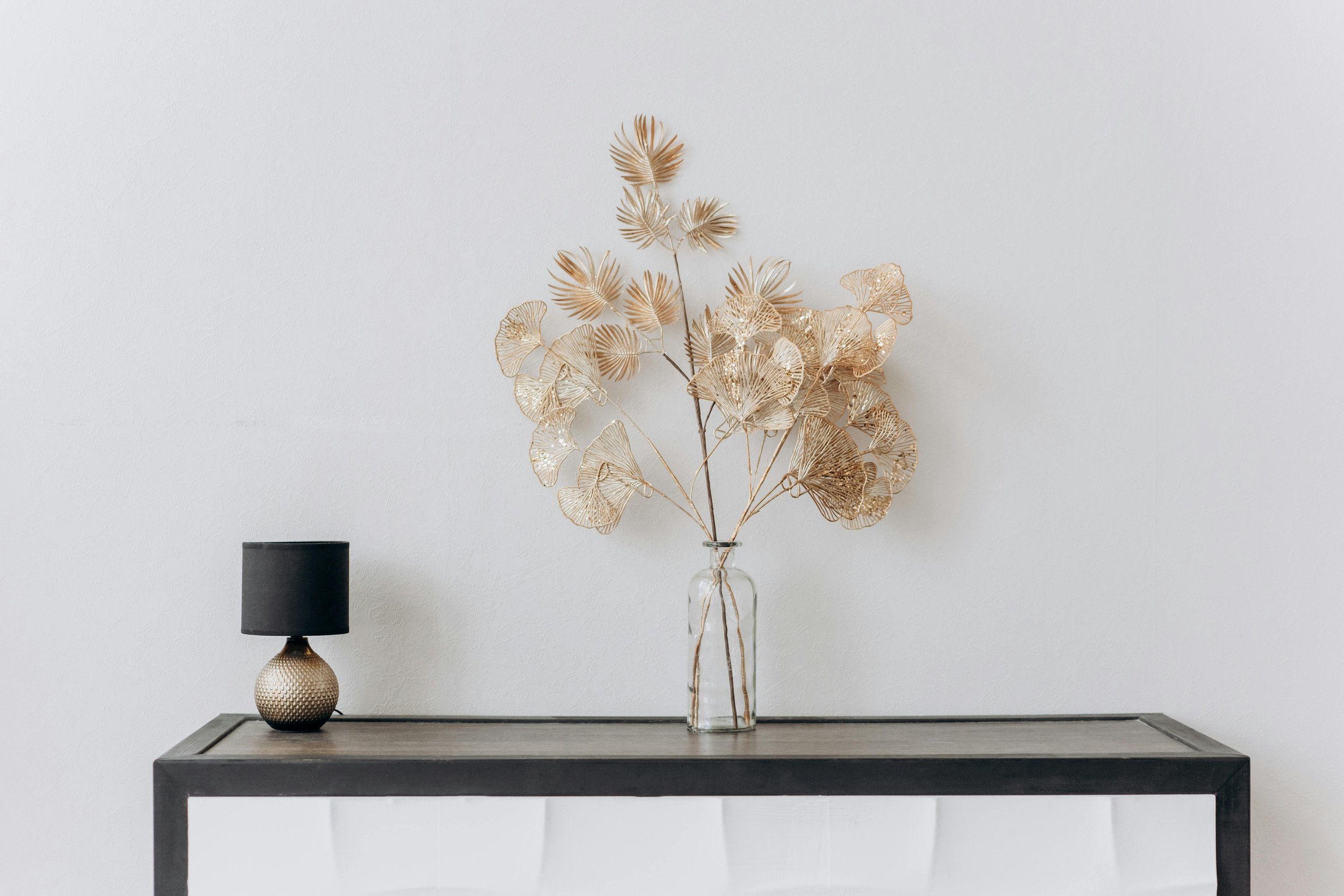 A modern minimalist interior displaying a wooden table with a glass vase holding dried palm and fan-shaped leaves, a textured gold table lamp with a black shade, and a white wall background.