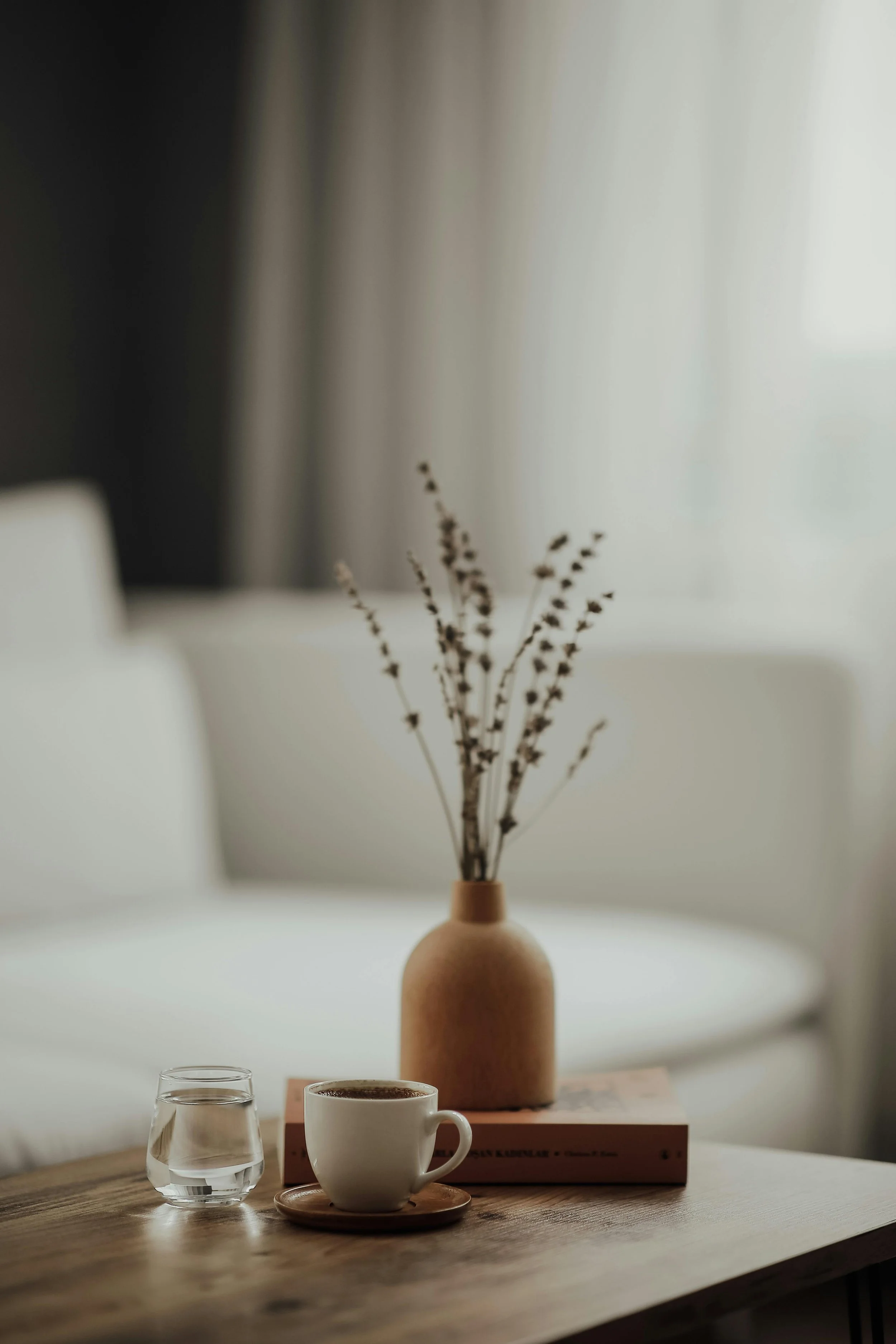 A cozy table setup with a clear glass of water, a white coffee cup on a small wooden plate, and a brown book underneath a tall, beige vase holding dried branches, all on a wooden table in a softly lit room.