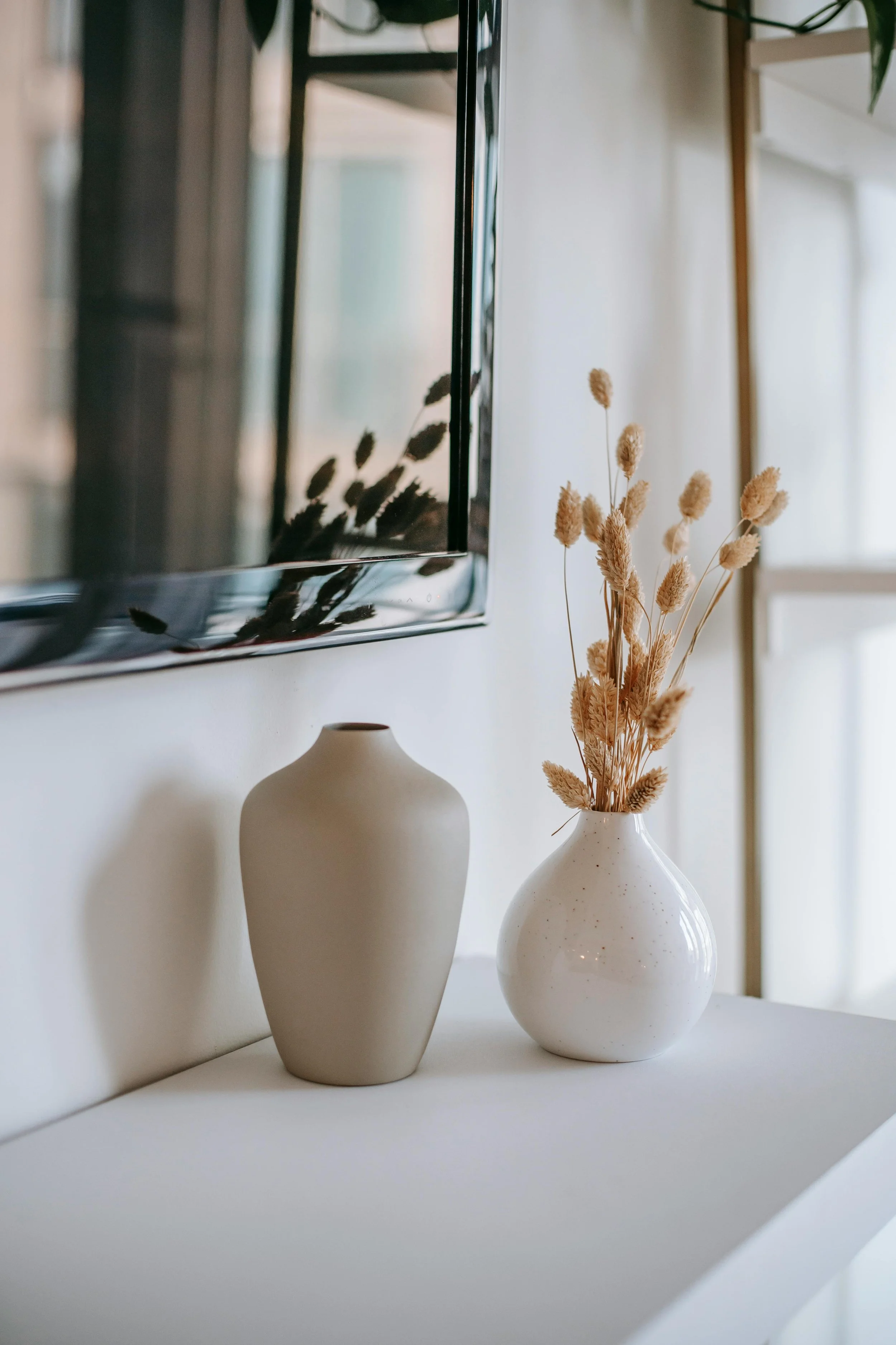 Decorative interior scene with two white ceramic vases, one filled with dried beige wheat stalks, placed on a white surface with a wall and mirror reflecting dried wheat stalks in the background.