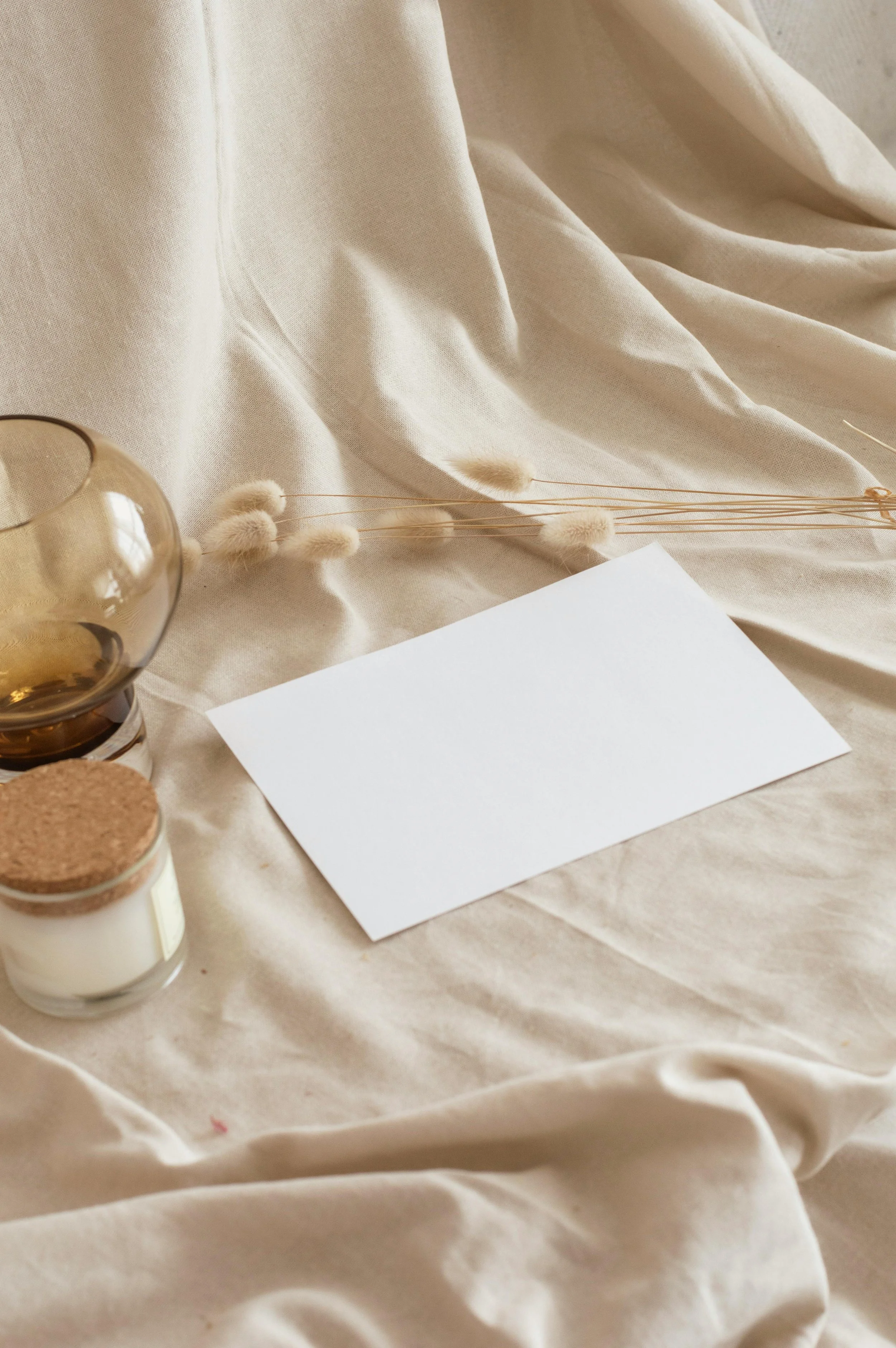 Cream-colored fabric, dried flowers, a glass container, a white candle, a corked glass jar, and a blank white paper on a soft beige surface.