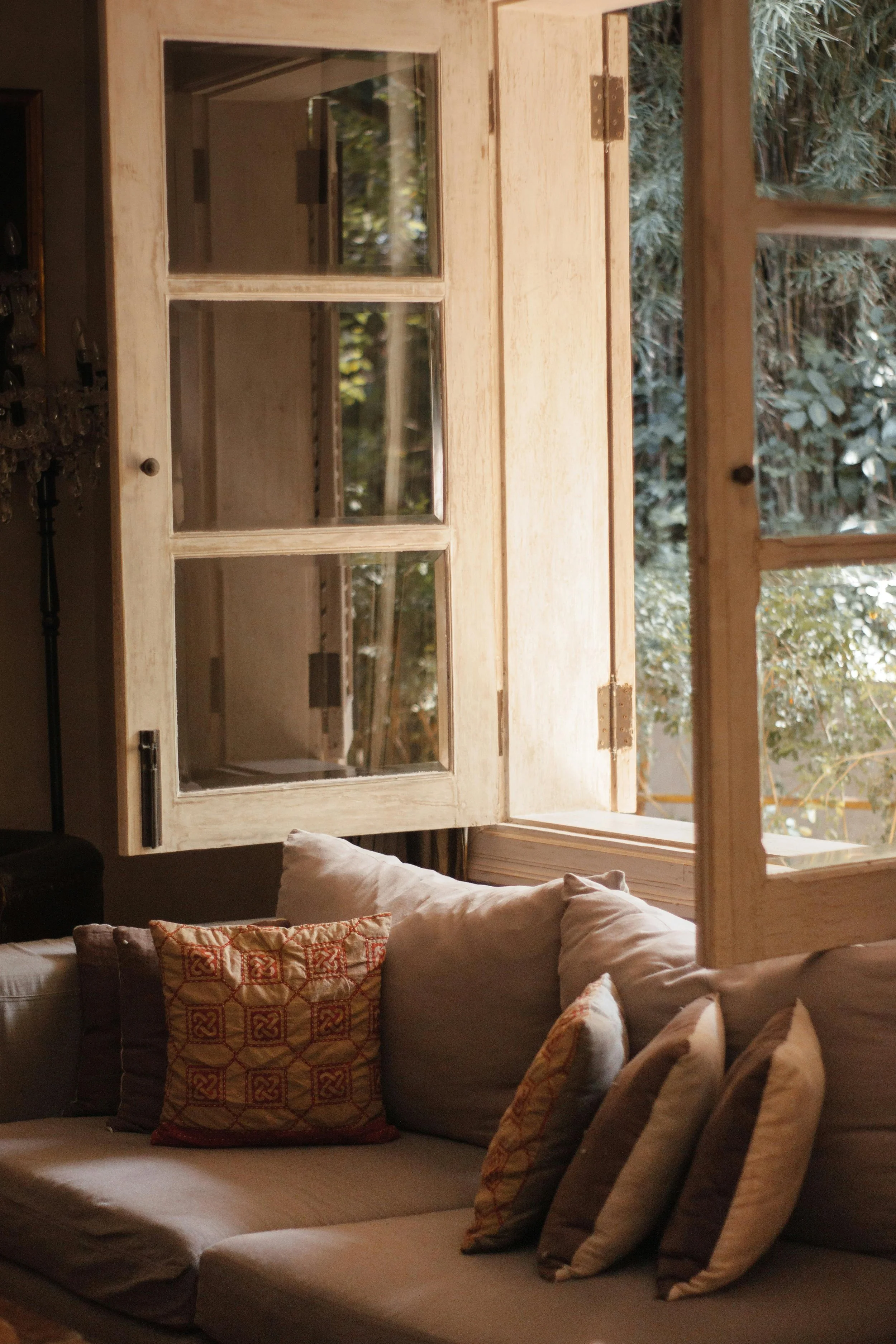 A cozy living room with a beige sofa adorned with multiple decorative pillows, positioned in front of large wooden-framed windows revealing a lush outdoor garden with greenery and trees. Sunlight softly illuminates the space, creating a warm atmosphere.