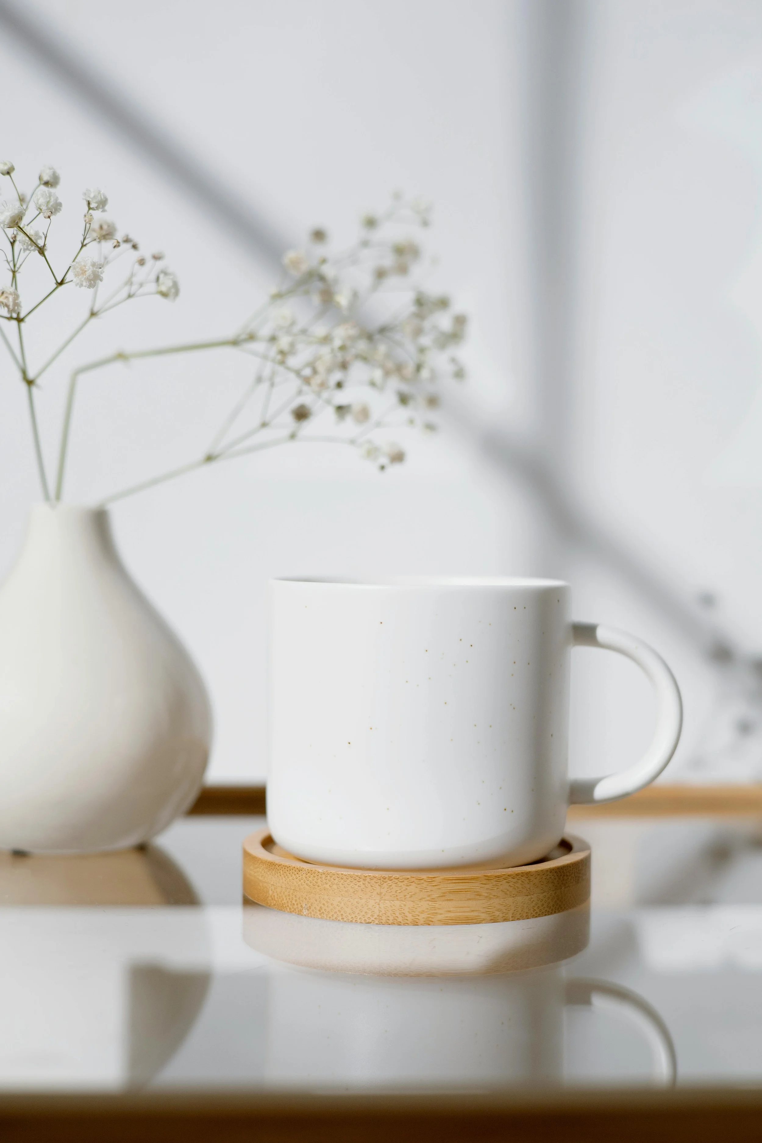 A white ceramic mug on a round wooden coaster, with a white vase of small white flowers in the background.