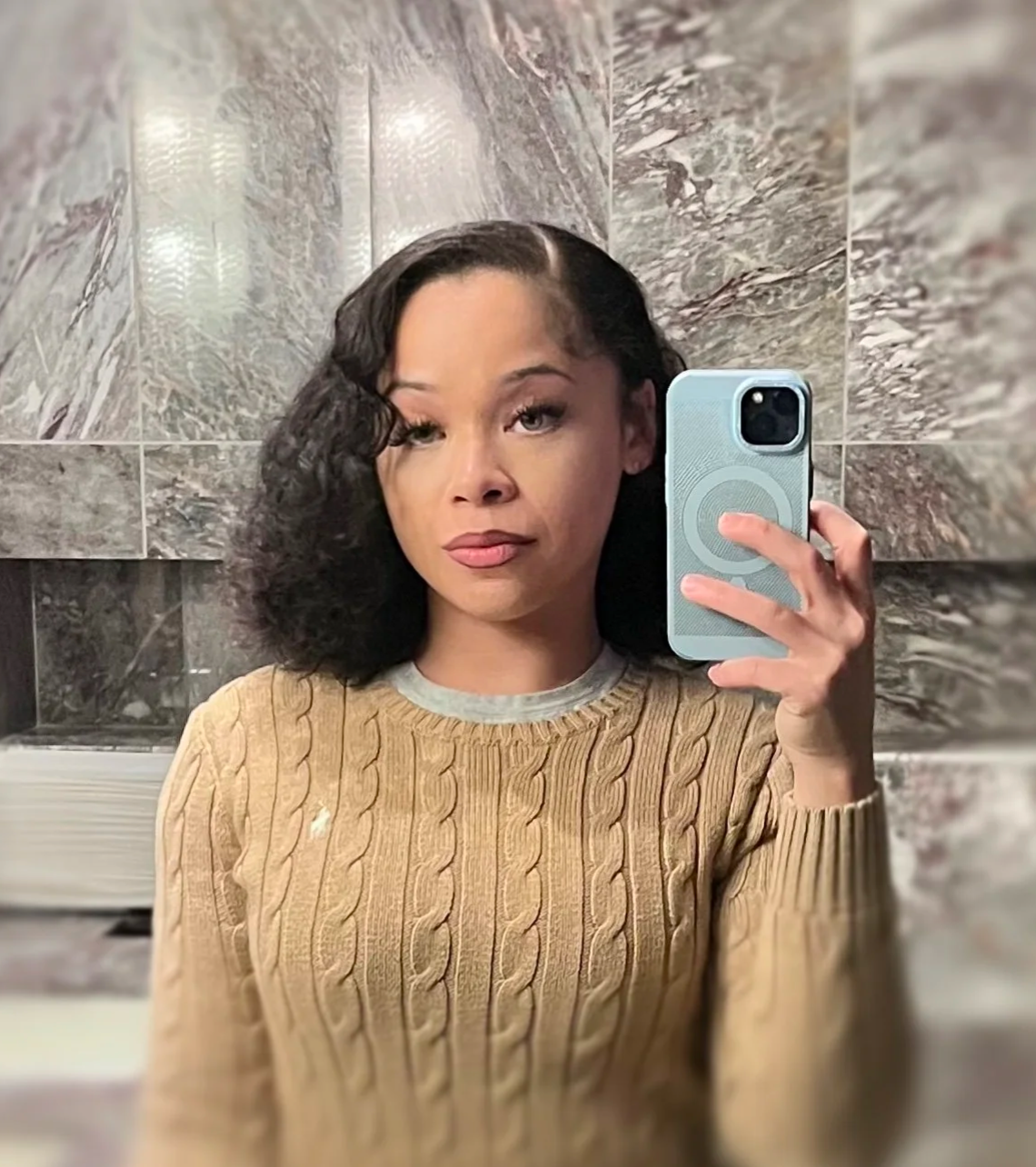 A woman taking a selfie in a bathroom mirror. She has curly black hair, light brown skin, and is wearing a beige cable-knit sweater.