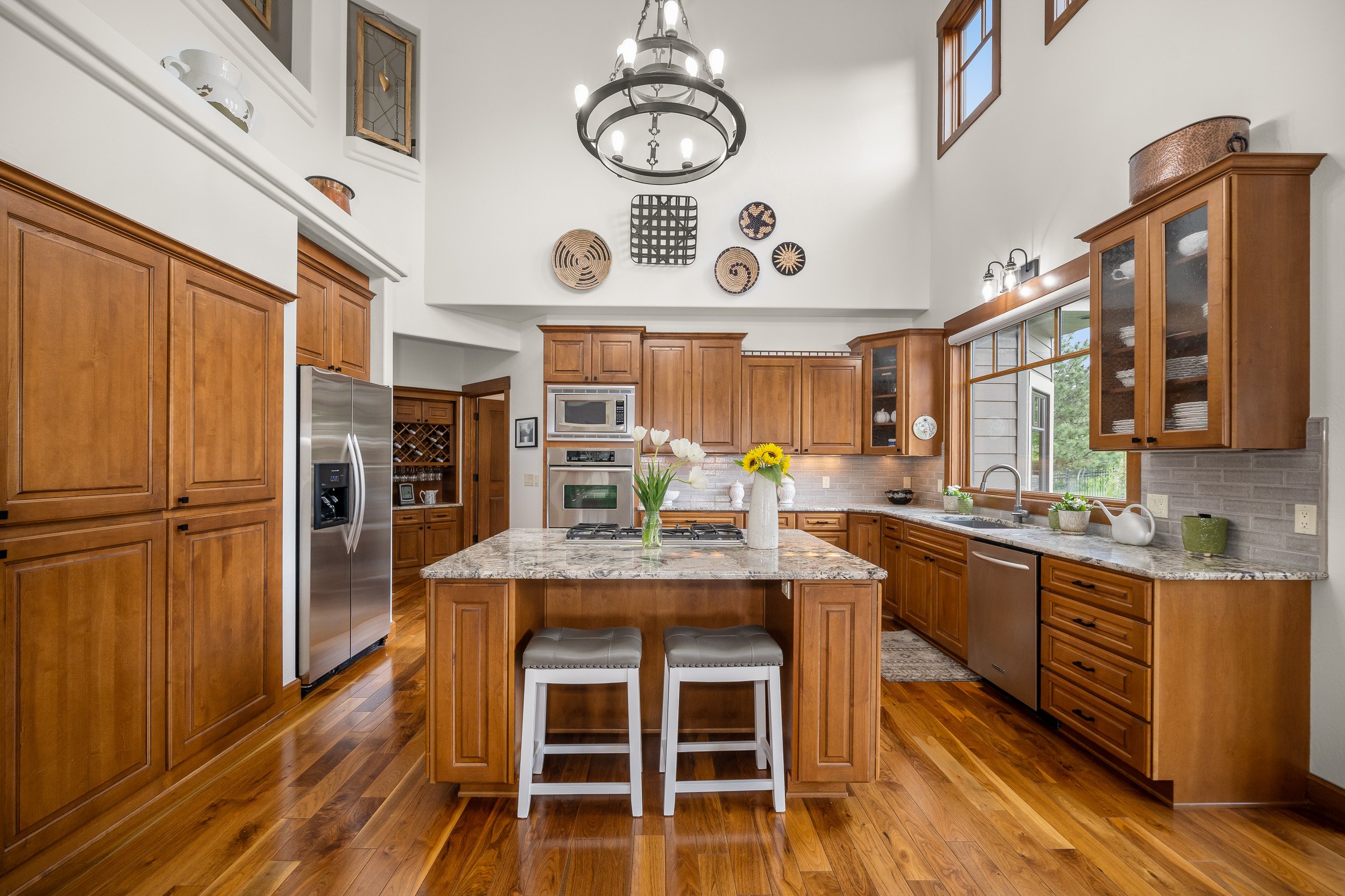 A spacious kitchen with wooden cabinets, a central island with a granite countertop, stainless steel appliances, a vase with flowers, and decorative wall art under a large chandelier.